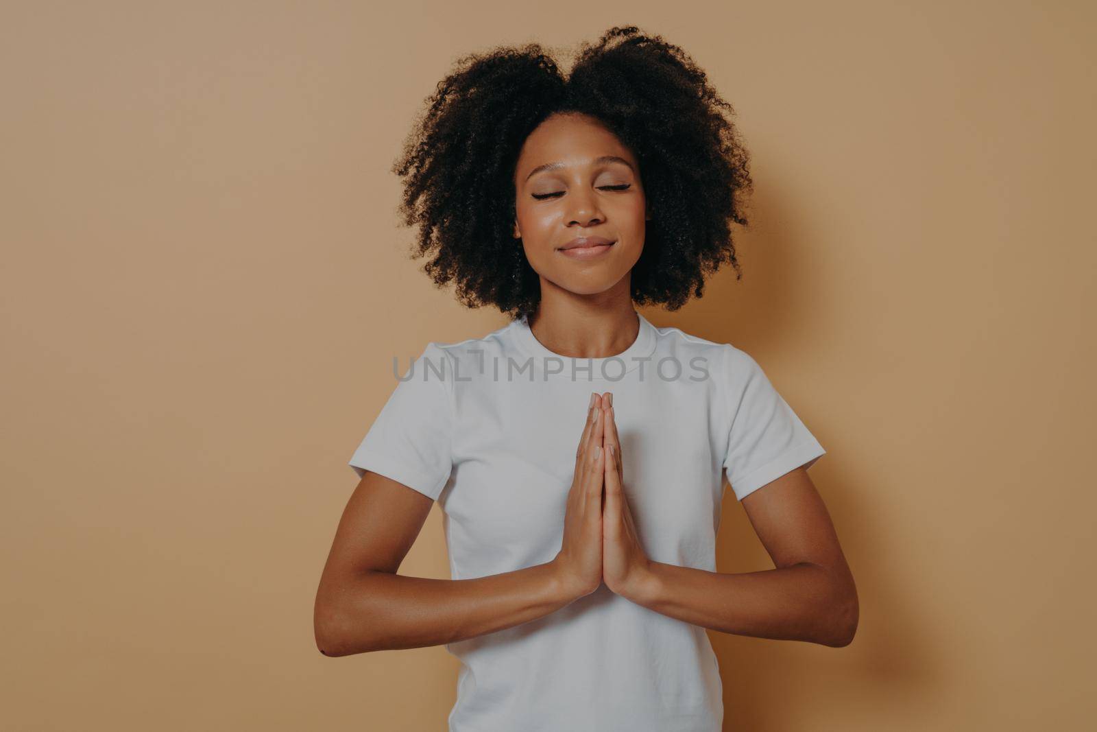 African american young woman holding hands clasped together and praying on beige background by vkstock