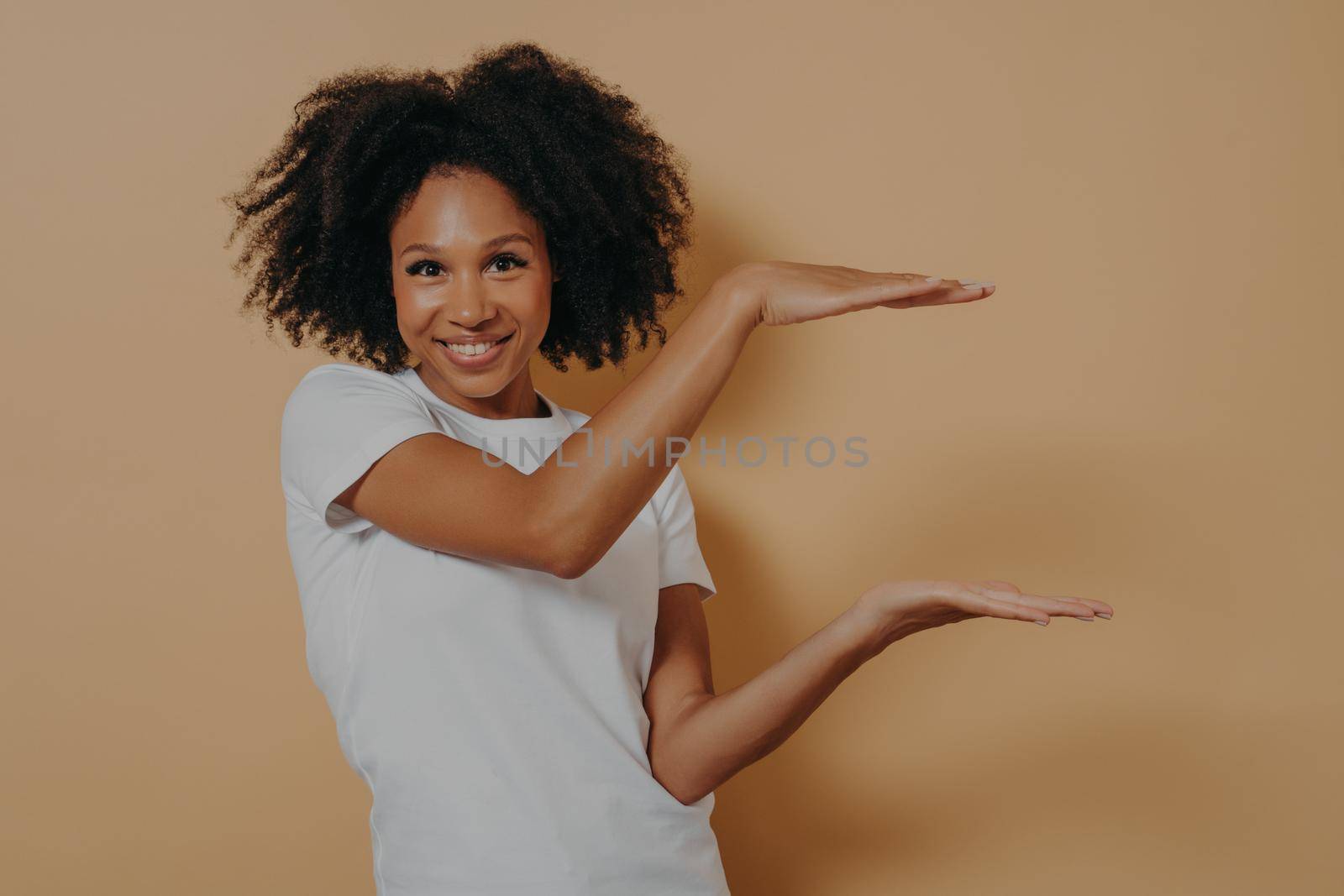 Young dark skinned showing large object, shaping box with both hands, isolated over beige background by vkstock