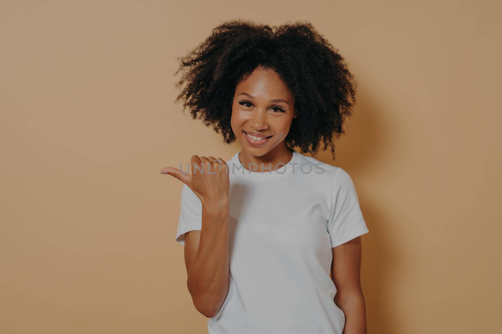 Young smiling dark skinned female curly hair pointing aside with thumb, shows blank space for promotion or advertisement with positive face expression, isolated over beige wall
