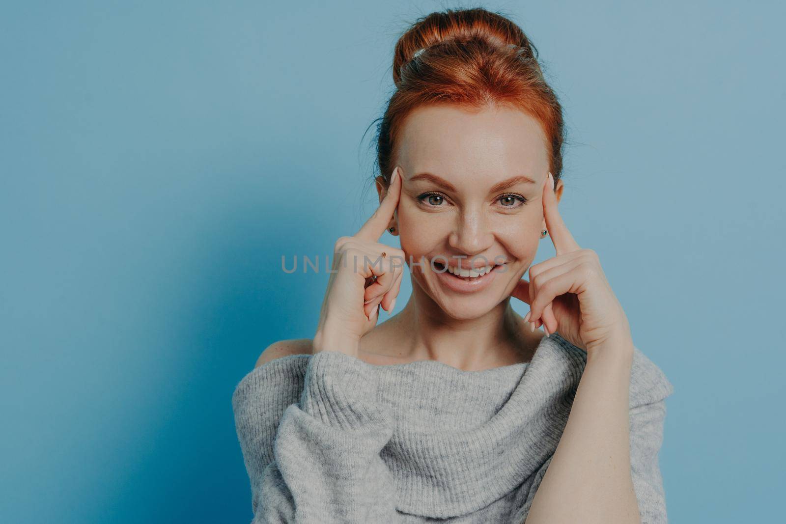 Headshot of young positive red haired woman with fingers on temples smiling at camera while standing isolated on blue background, thinking and trying to focus on task, dressed in knitted sweater