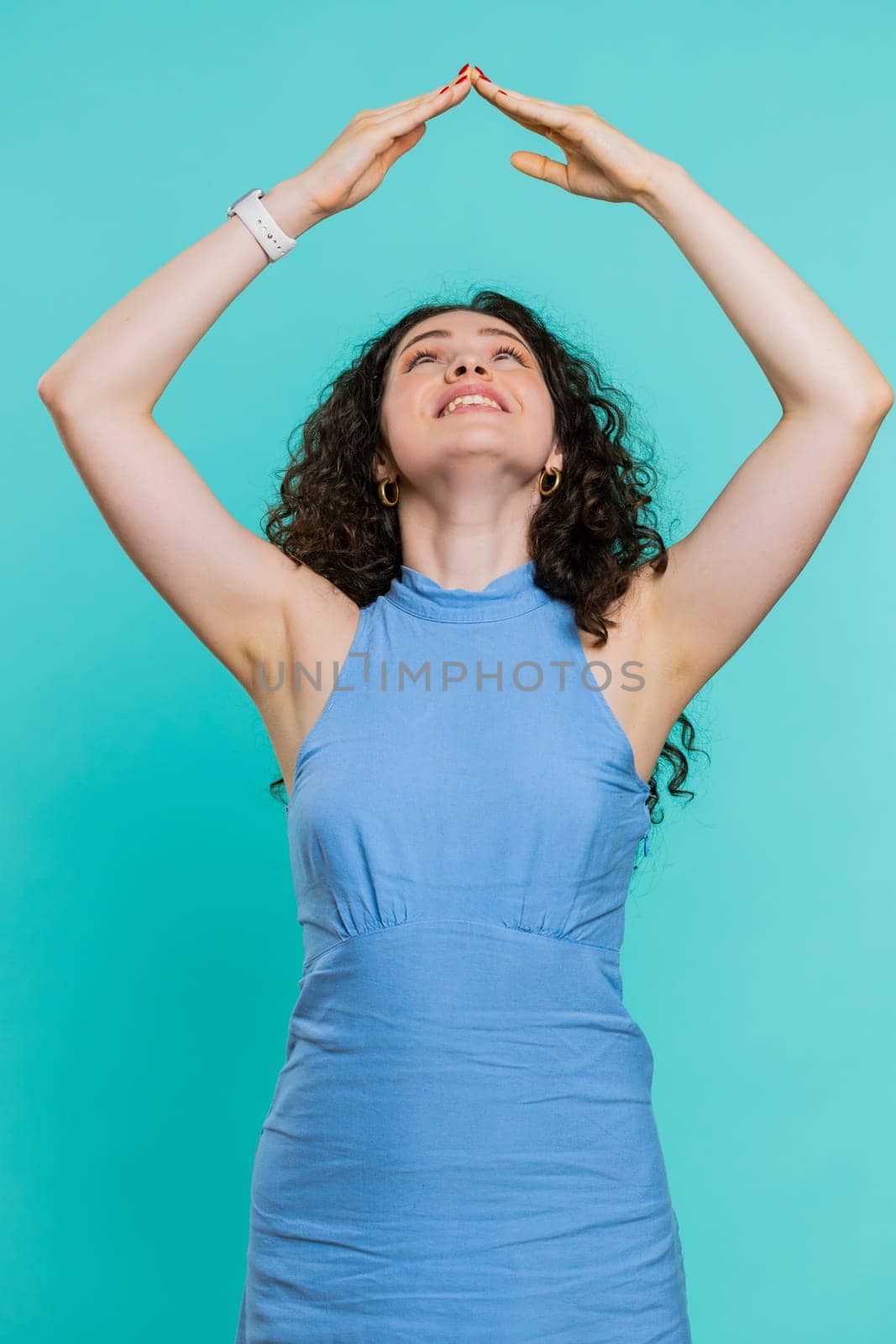 House roof about head. Young woman playing childish catching up game, feeling in safe making roof above head with hands, insurance, security service. Girl isolated on blue studio background. Vertical