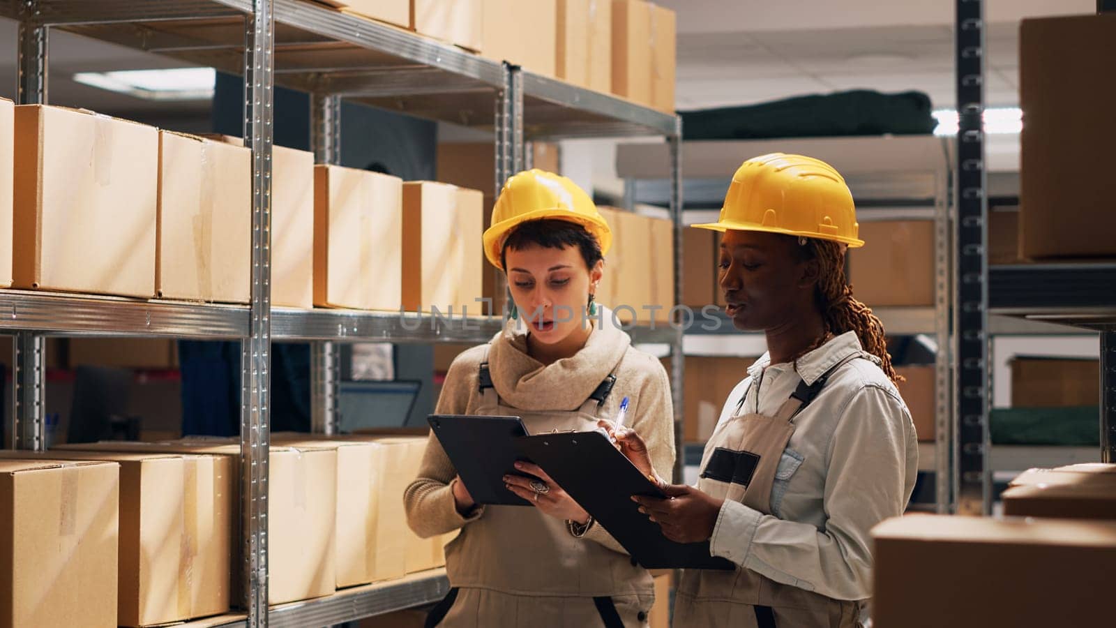 Diverse employees counting boxes of products placed on storehouse shelves, looking at merchandise goods in cardboard packages. Two people in overalls working with cargo in warehouse depot.