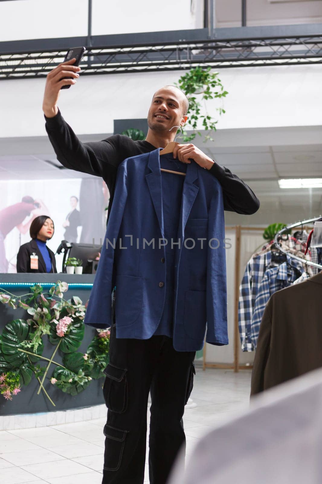 Clothing store arab man client holding jacket on hanger and taking photo on smartphone while browsing apparel in shopping mall. Boutique smiling buyer chatting in videocall and showing outfit