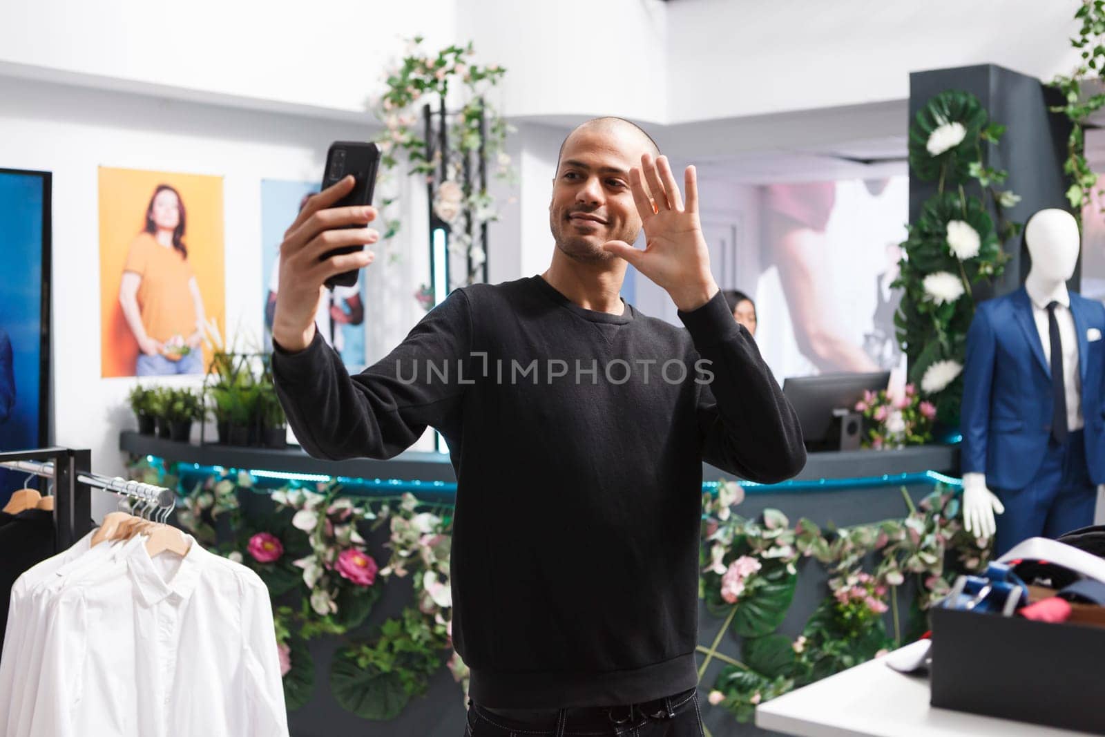 Young smiling arab man taking selfie on smartphone in clothing store to post on fashion blog. Boutique buyer using mobile phone while greeting online channel follower and looking at front camera