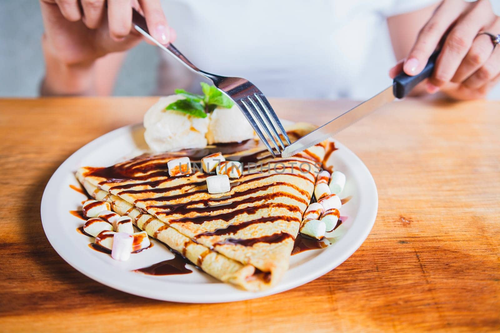 Hands of person with fork cutting chocolate crepe and ice cream. Woman eating chocolate crepe and ice cream with fork by isaiphoto