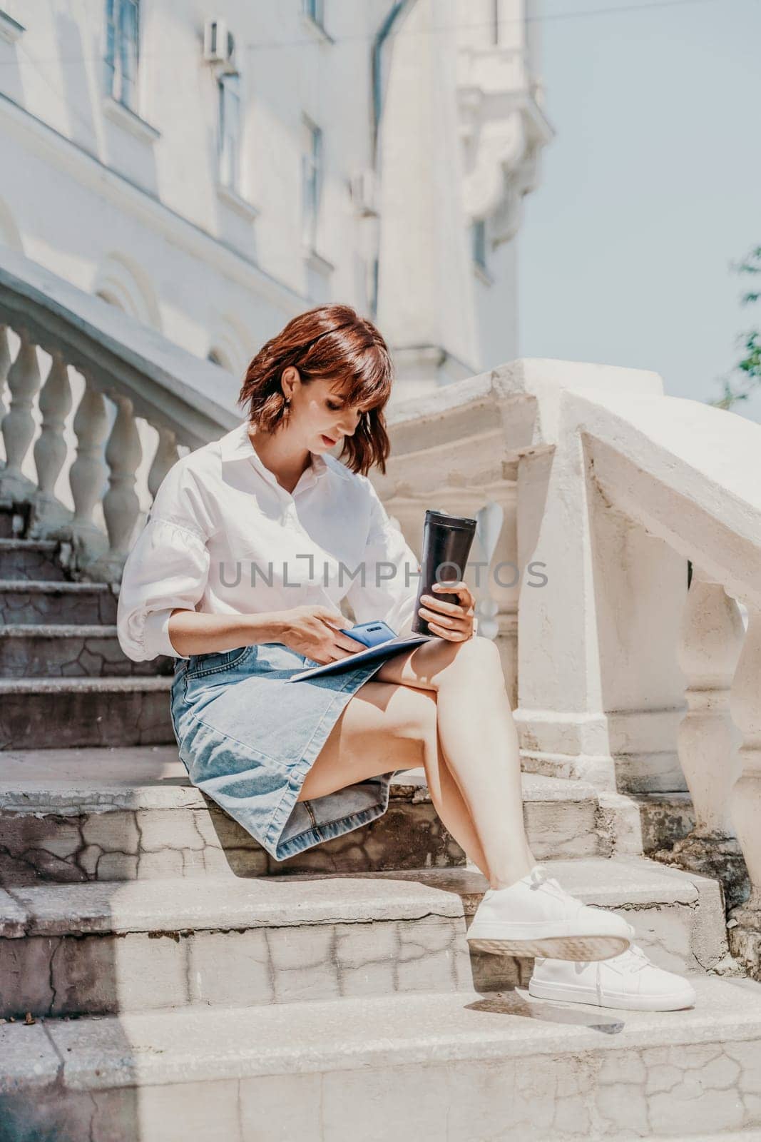 Freelancer woman phone. A business woman in a white shirt and denim skirt sits on the steps near an ancient building in the city and looks into the phone.