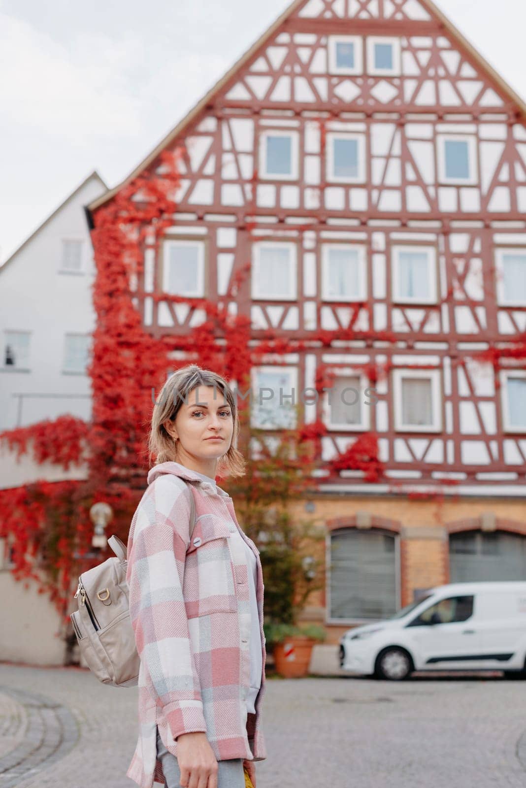 a beautiful girl stands against the background of the window of an old European house, entwined with a floating red color in autumn. Tourism & Travel Concept. Nice portrait of a young woman, in boho style outdoors in fall autumn day. Caucasian female girl 20 30 years old