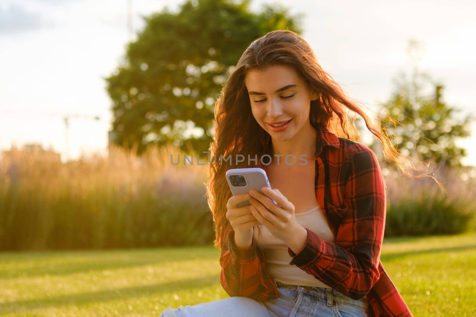 The happy millennial girl checking social media holding a smartphone sitting on the grass in the park at sunset.