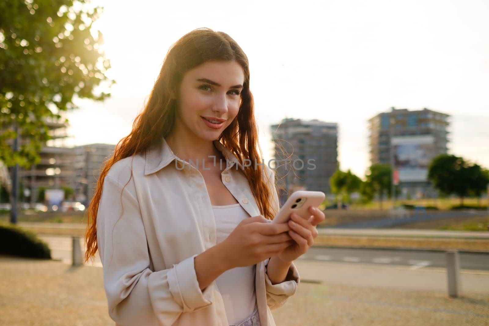 Portrait of red haired girl in casual clothes standing with a mobile phone in the street.