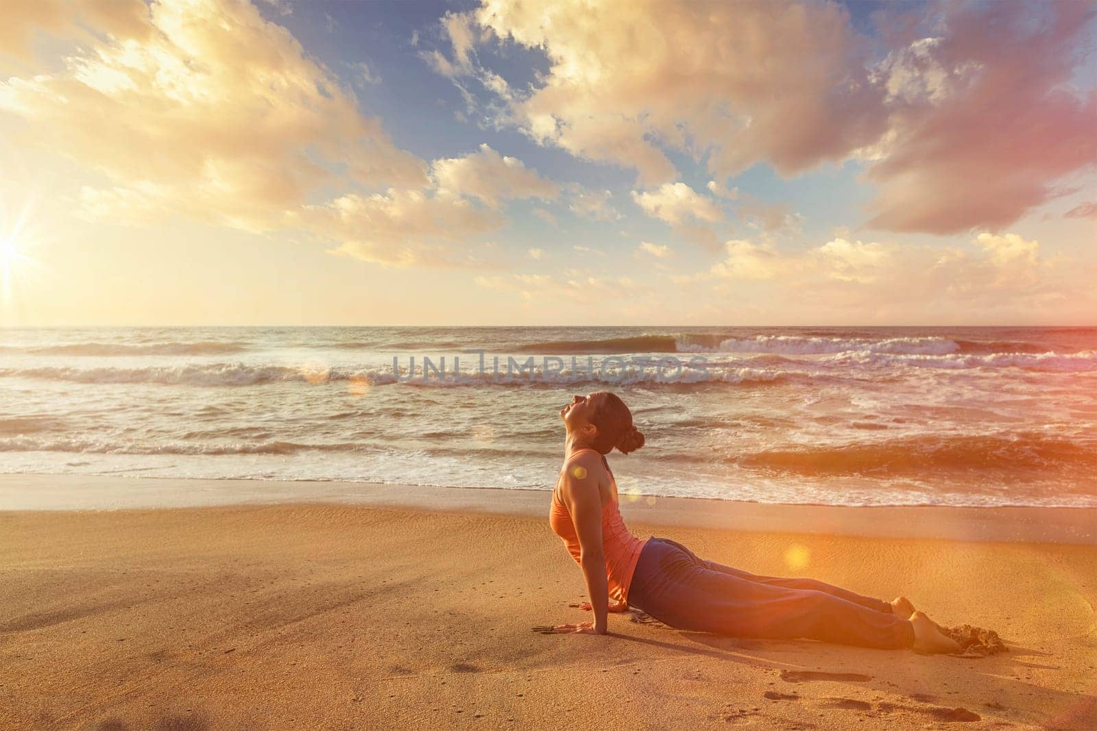 Yoga outdoors on beach - woman practices Ashtanga Vinyasa yoga Surya Namaskar Sun Salutation asana Urdhva Mukha Svanasana - upward facing dog pose on sunset. With light leak and lens flare