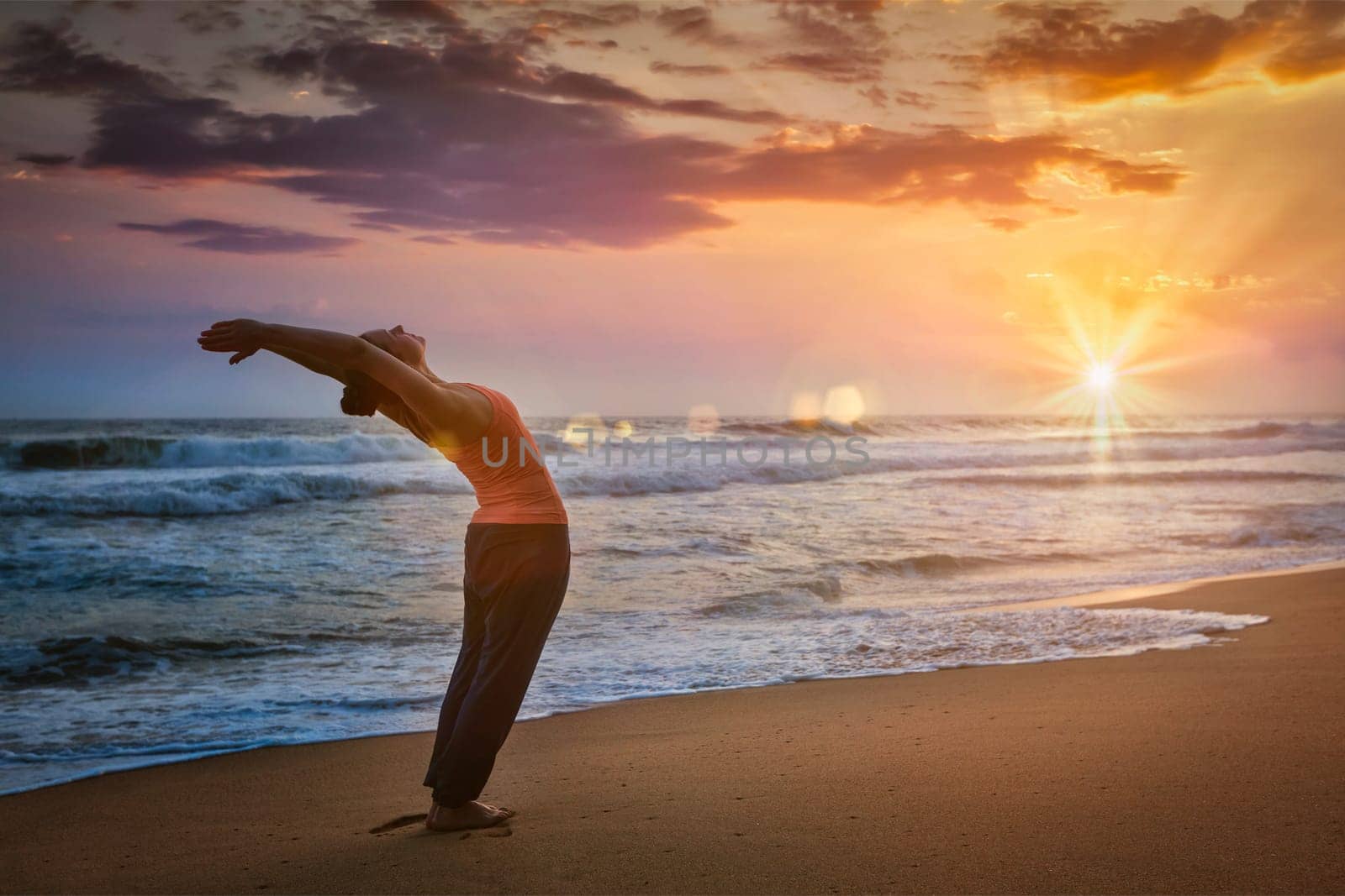 Young sporty fit woman doing yoga Sun salutation Surya Namaskar pose Hasta Uttanasana on tropical beach on sunset. With lens flare