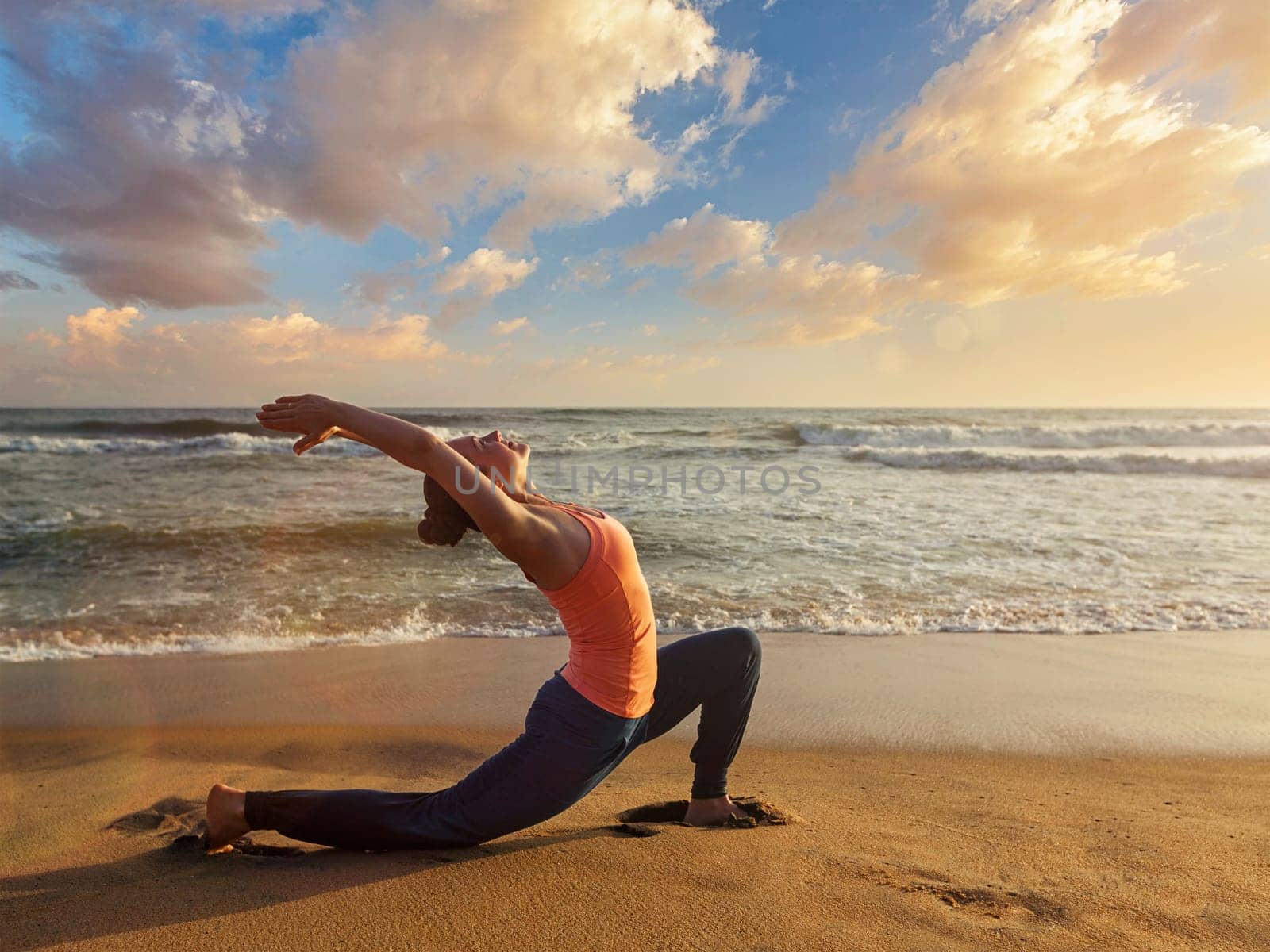 Sporty fit woman practices yoga Anjaneyasana at beach on sunset by dimol