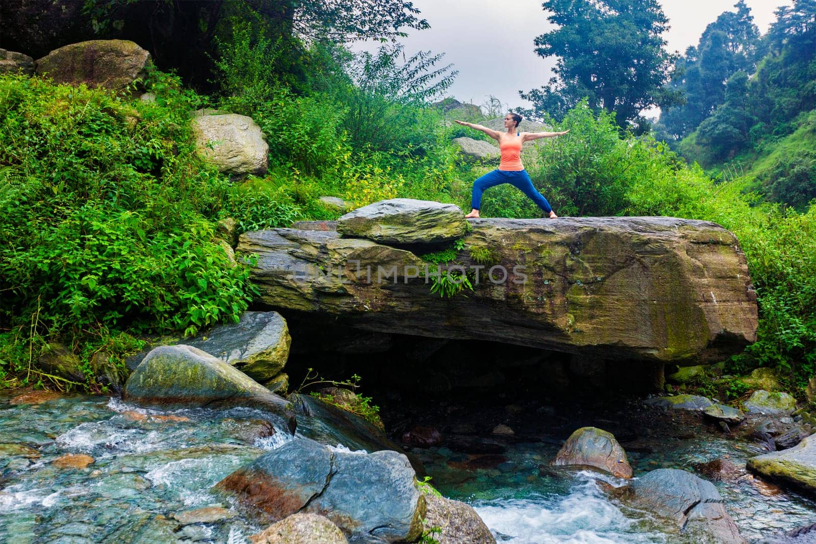 Yoga outdoors - sporty fit woman doing Ashtanga Vinyasa Yoga asana Virabhadrasana 2 Warrior pose posture at tropical waterfall