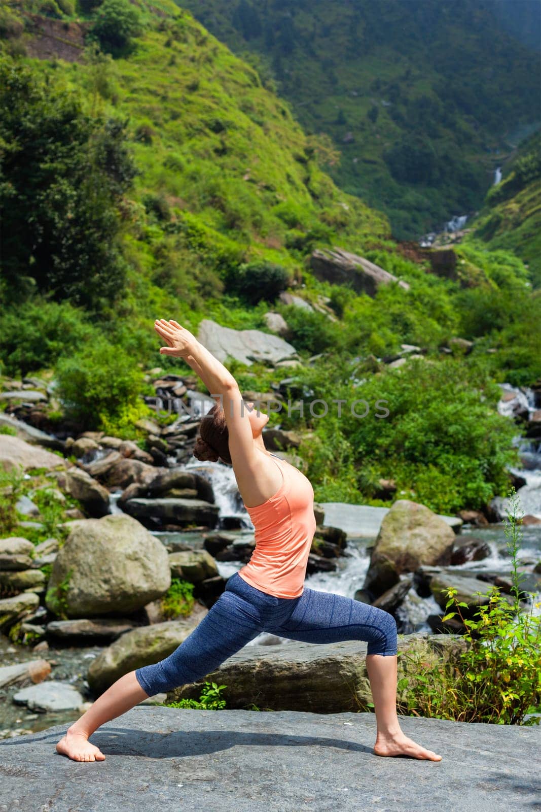 Yoga outdoors - sporty fit woman doing Ashtanga Vinyasa Yoga asana Virabhadrasana 1 Warrior pose posture at waterfall in HImalayas mountains