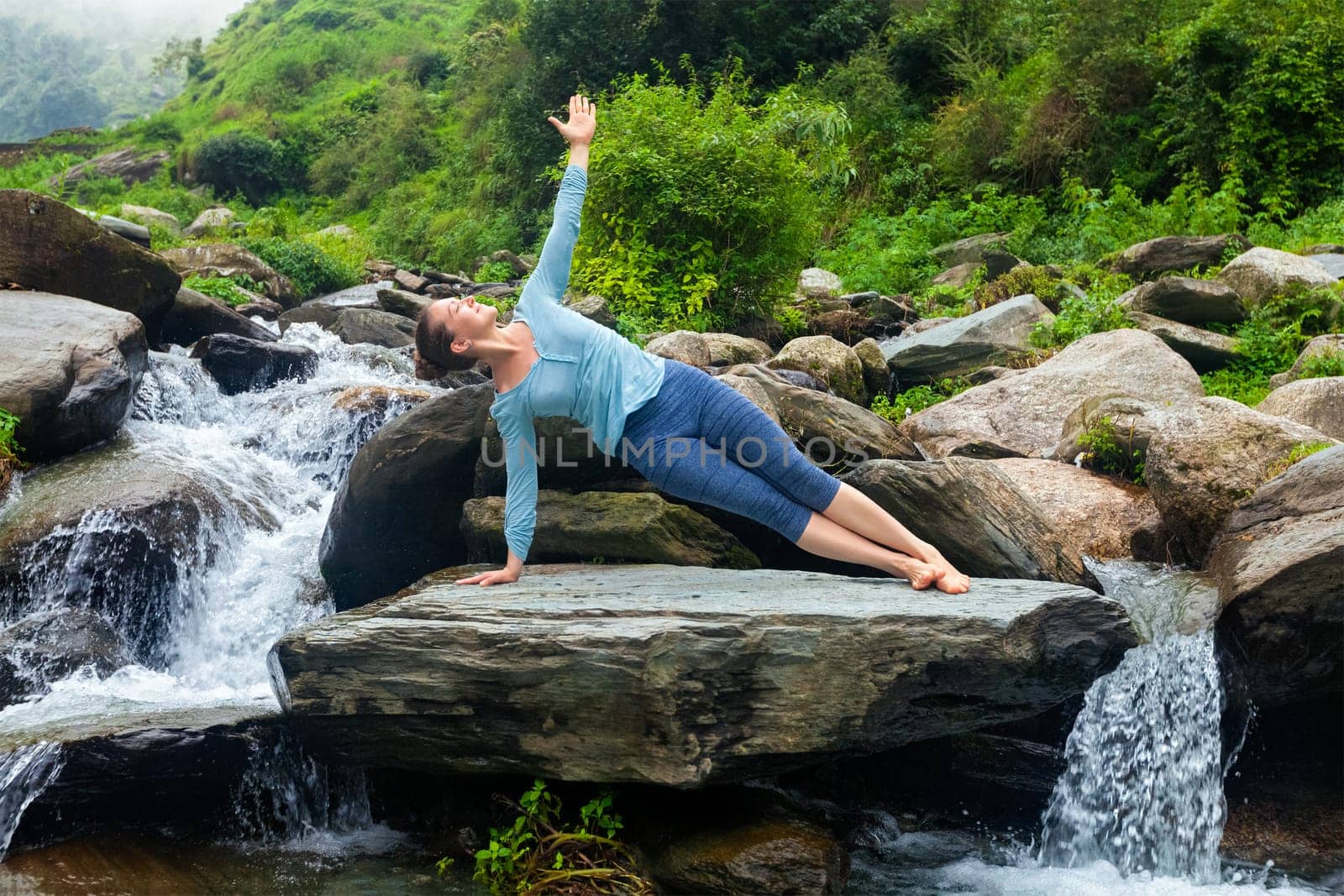 Yoga outdoors - beautiful sporty fit woman doing yoga asana Vasisthasana - side plank pose at tropical waterfall