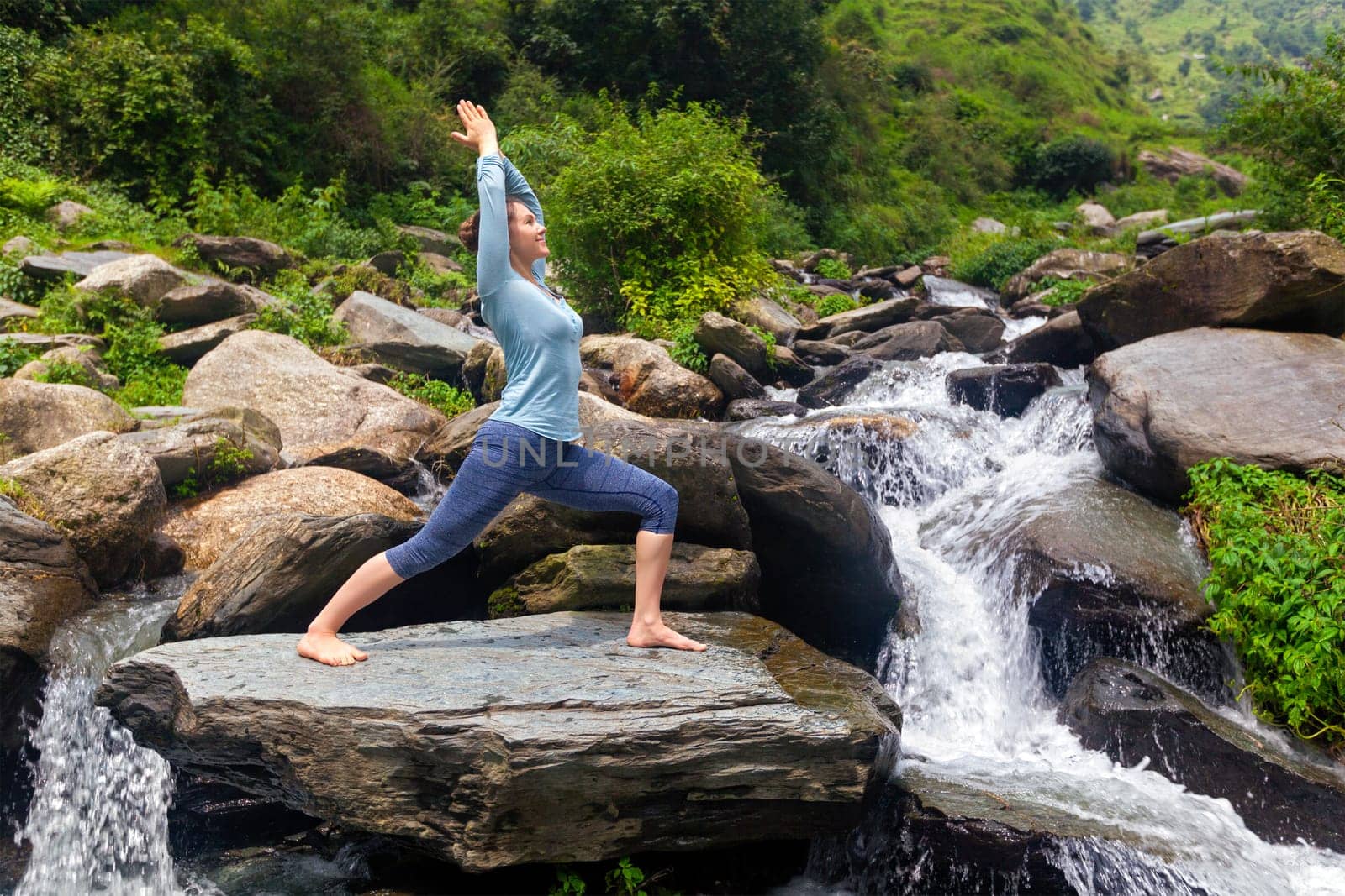 Yoga outdoors - sporty fit woman doing Ashtanga Vinyasa Yoga asana Virabhadrasana 1 Warrior pose posture at waterfall in HImalayas mountains