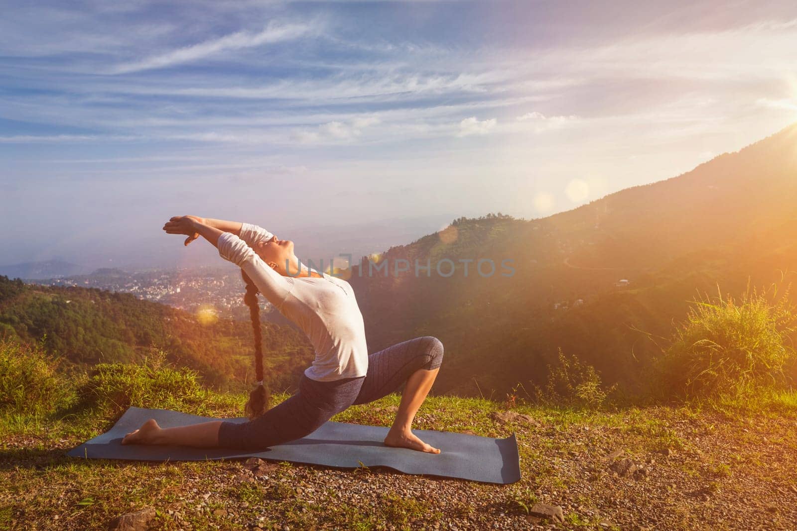 Yoga outdoors - sporty fit woman practices yoga Anjaneyasana - low crescent lunge pose outdoors in mountains in morning. With light leak and lens flare