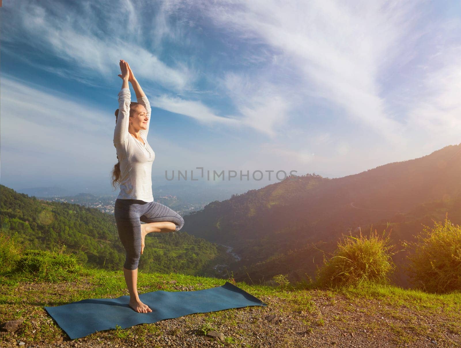 Yoga practice outdoors - woman practices balance yoga asana Vrikshasana tree pose in Himalayas mountains outdoors in the morning. Himachal Pradesh, India