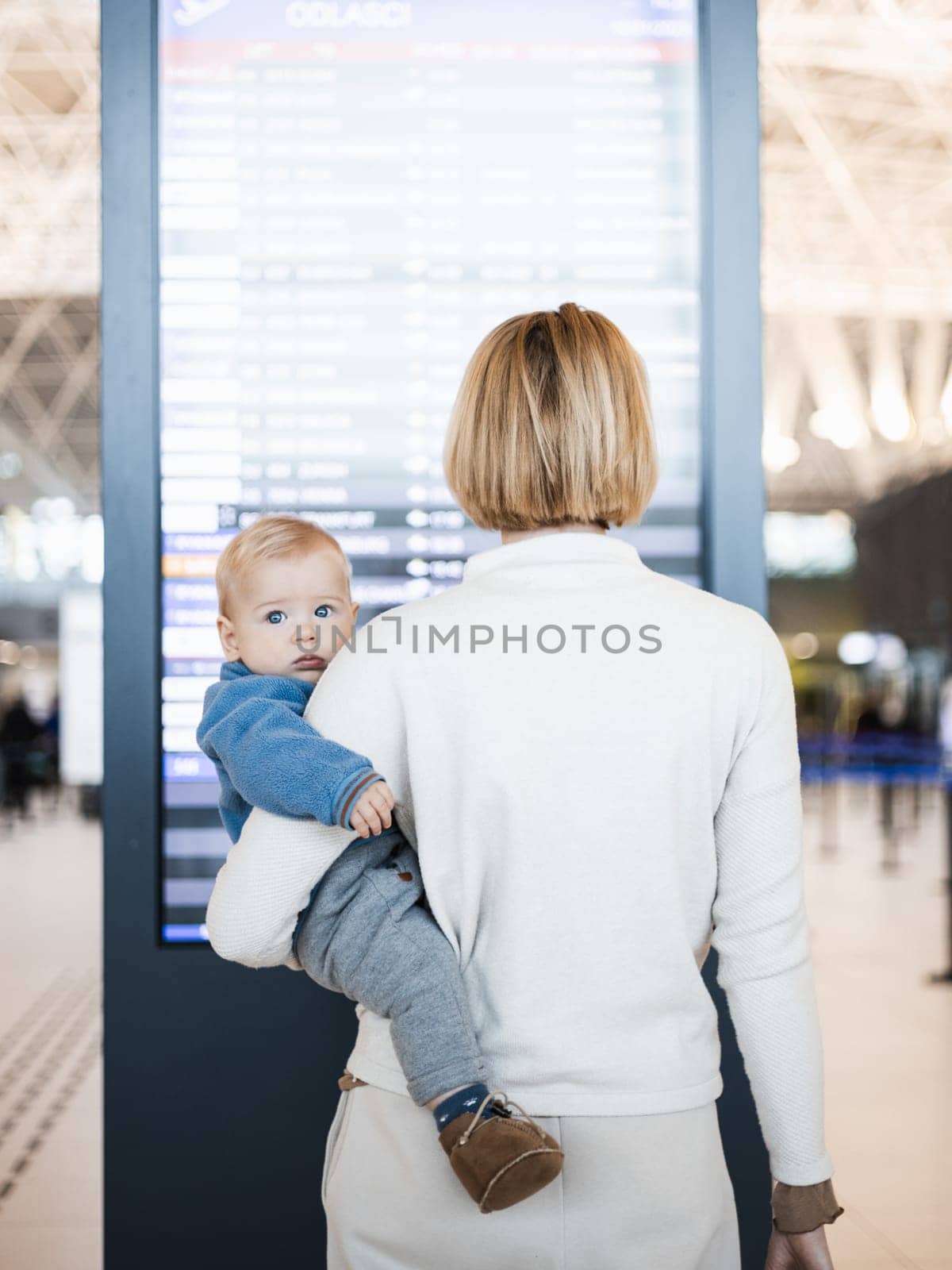 Mother traveling with child, holding his infant baby boy at airport terminal, checking flight schedule, waiting to board a plane. Travel with kids concept