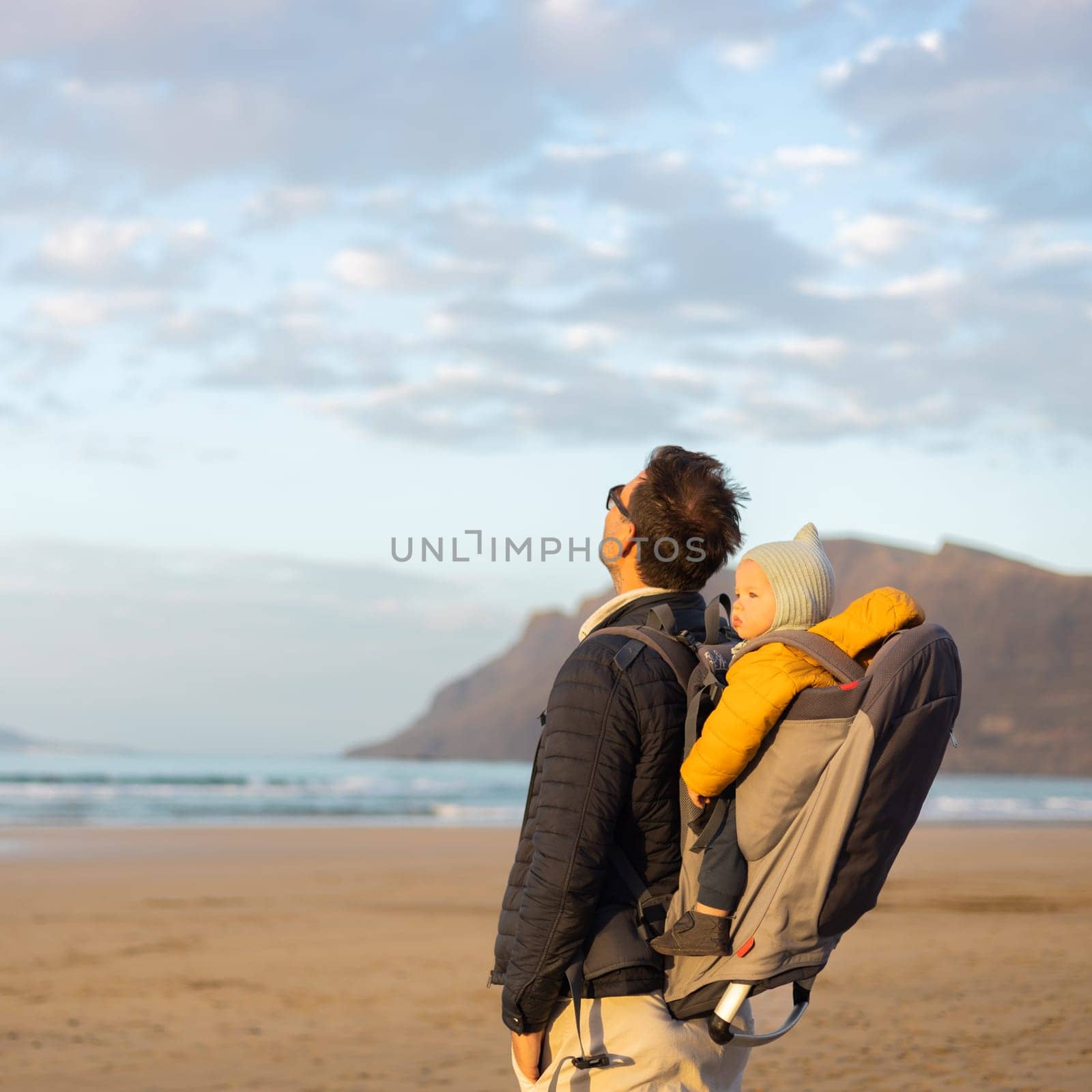 Father rising hands to the sky while enjoying pure nature carrying his infant baby boy son in backpack on windy sandy beach of Famara, Lanzarote island, Spain at sunset. Family travel concept