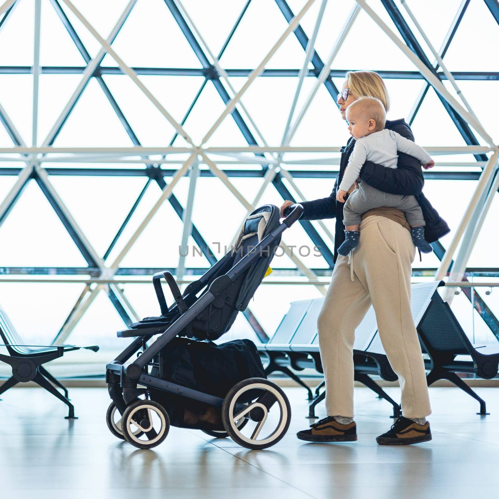 Mother carying his infant baby boy child, pushing stroller at airport departure terminal moving to boarding gates to board an airplane. Family travel with baby concept