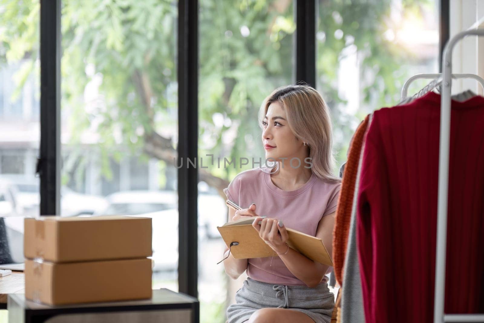 Female entrepreneur writes on parcel box after reading address on notebook. Online seller concept.