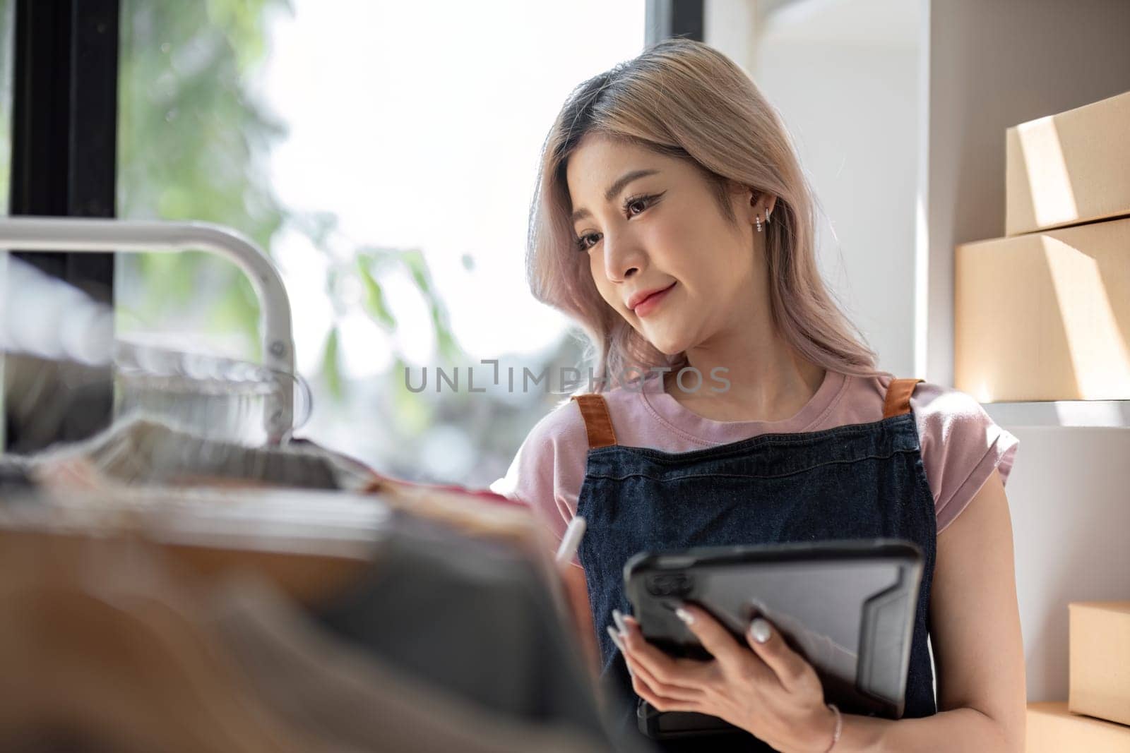 businesswoman owner an online trading business is using tablet to check the stock of the clothes on the rail rack at small office at home. work from home, online delivery, SME. by wichayada