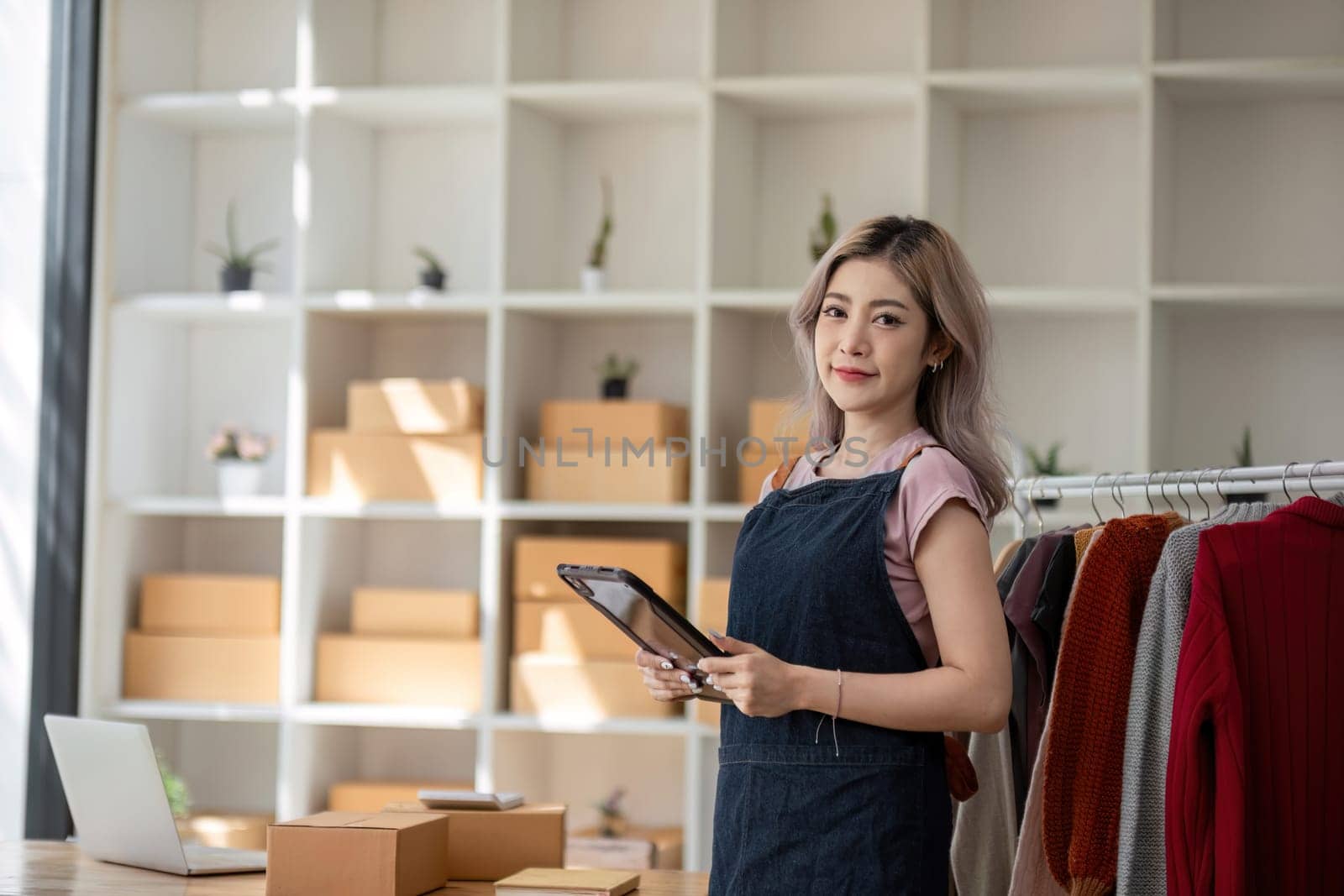 businesswoman owner an online trading business is using tablet to check the stock of the clothes on the rail rack at small office at home. work from home, online delivery, SME. by wichayada