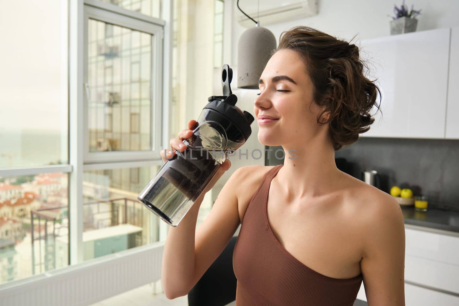 Sport and fitness concept. Portrait of young woman athlete drinking water from gym bottle, smiling with pleased face, wearing acrivewear, standing in kitchen by Benzoix