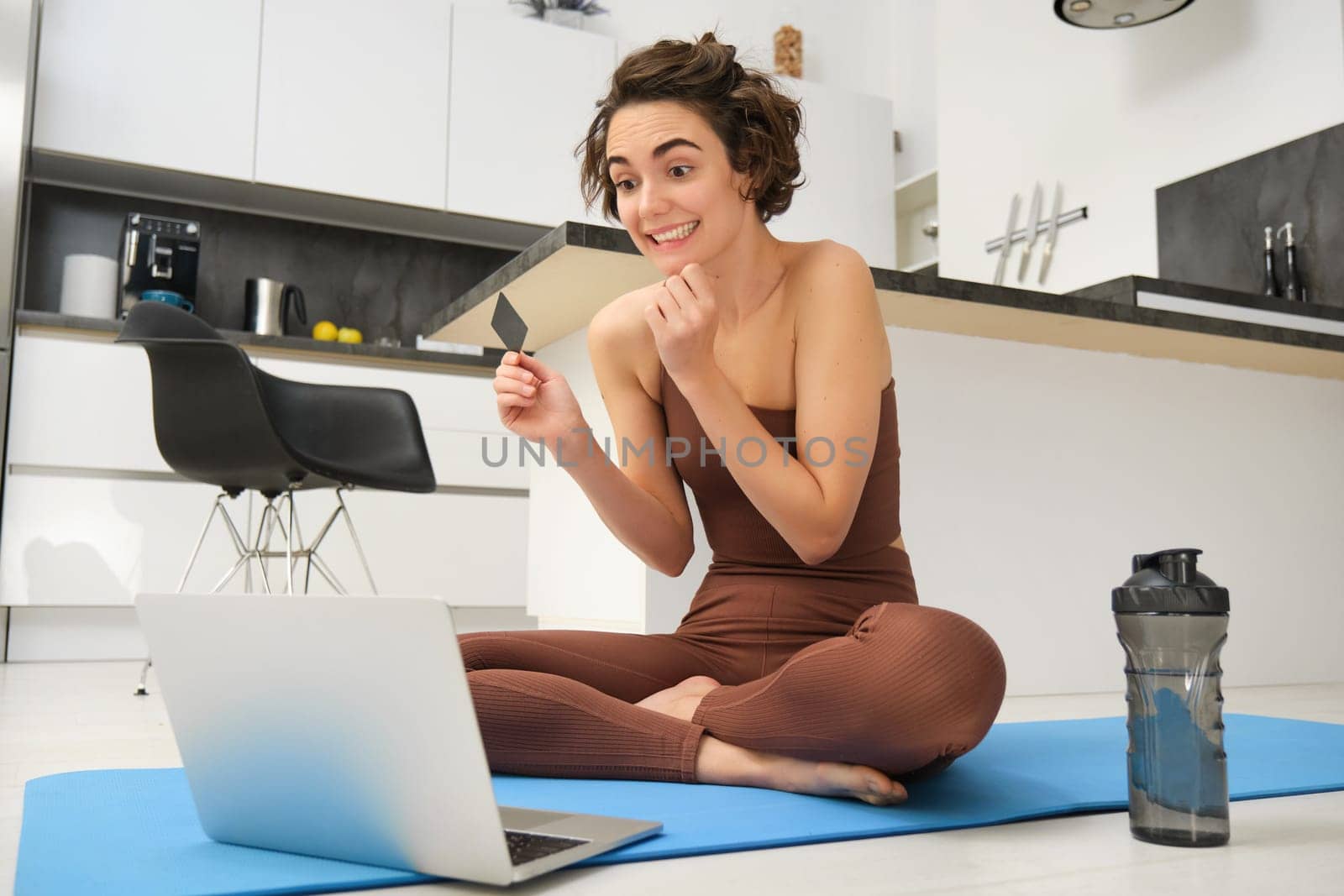 Portrait of young female athlete, yoga girl paying for online classes, purchasing remote training with gym instructor, sitting on her rubber mat with laptop and credit card.