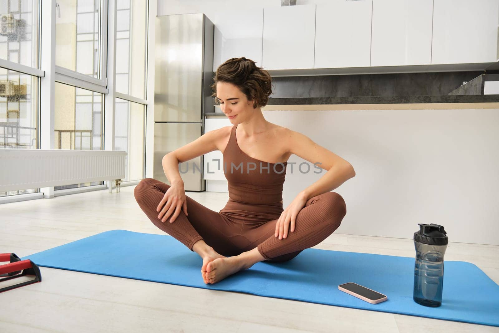 Portrait of fitness girl doing yoga on rubber mat at home, workout indoors in kitchen wearing sportswear, practice minfulness, sitting in lotus pose with water bottle next to her.