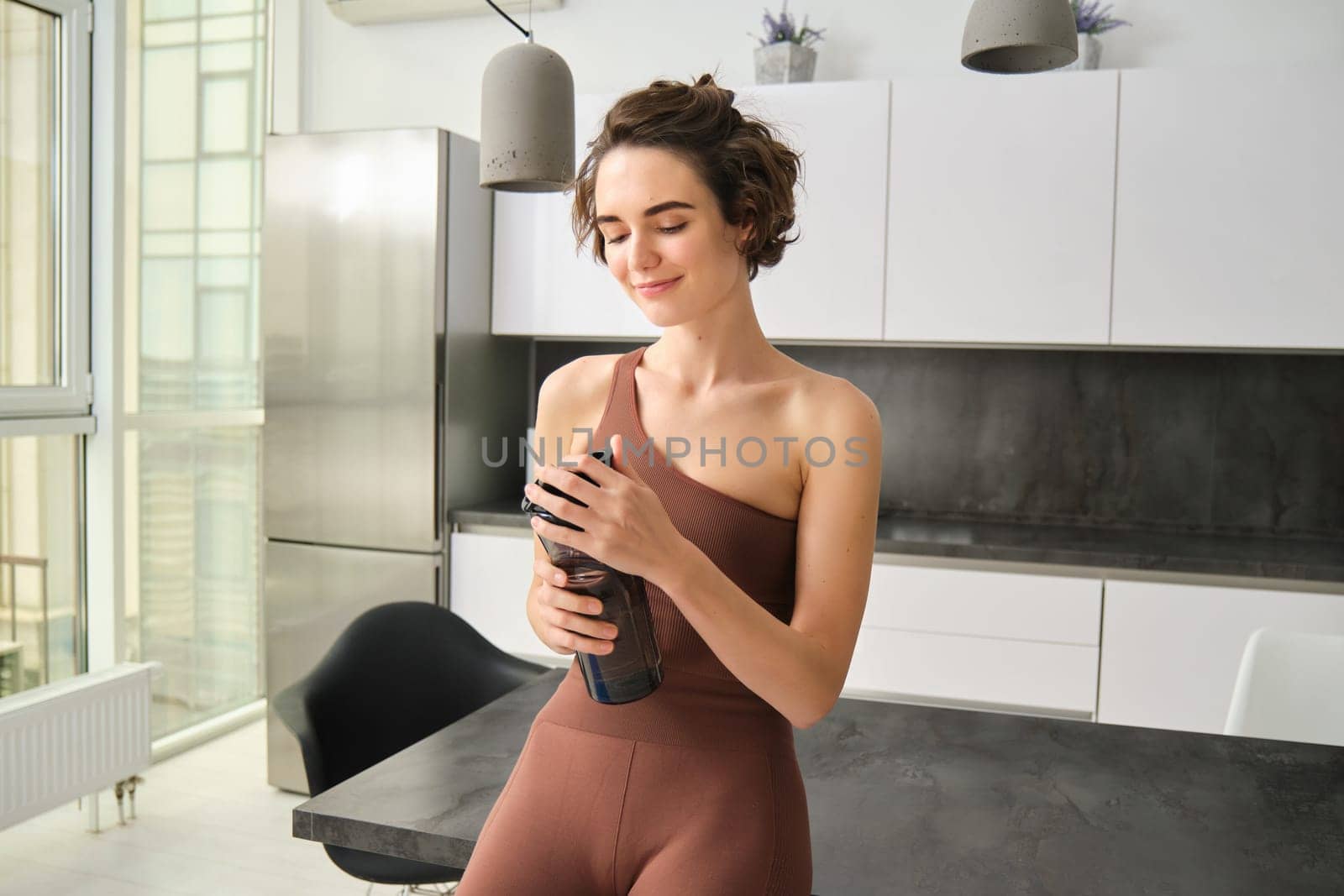 Sport and healthy lifestyle. Portrait of smiling fitness woman in activewear, standing near kitchen counter at home, drinking from water bottle, preparing for workout gym training.