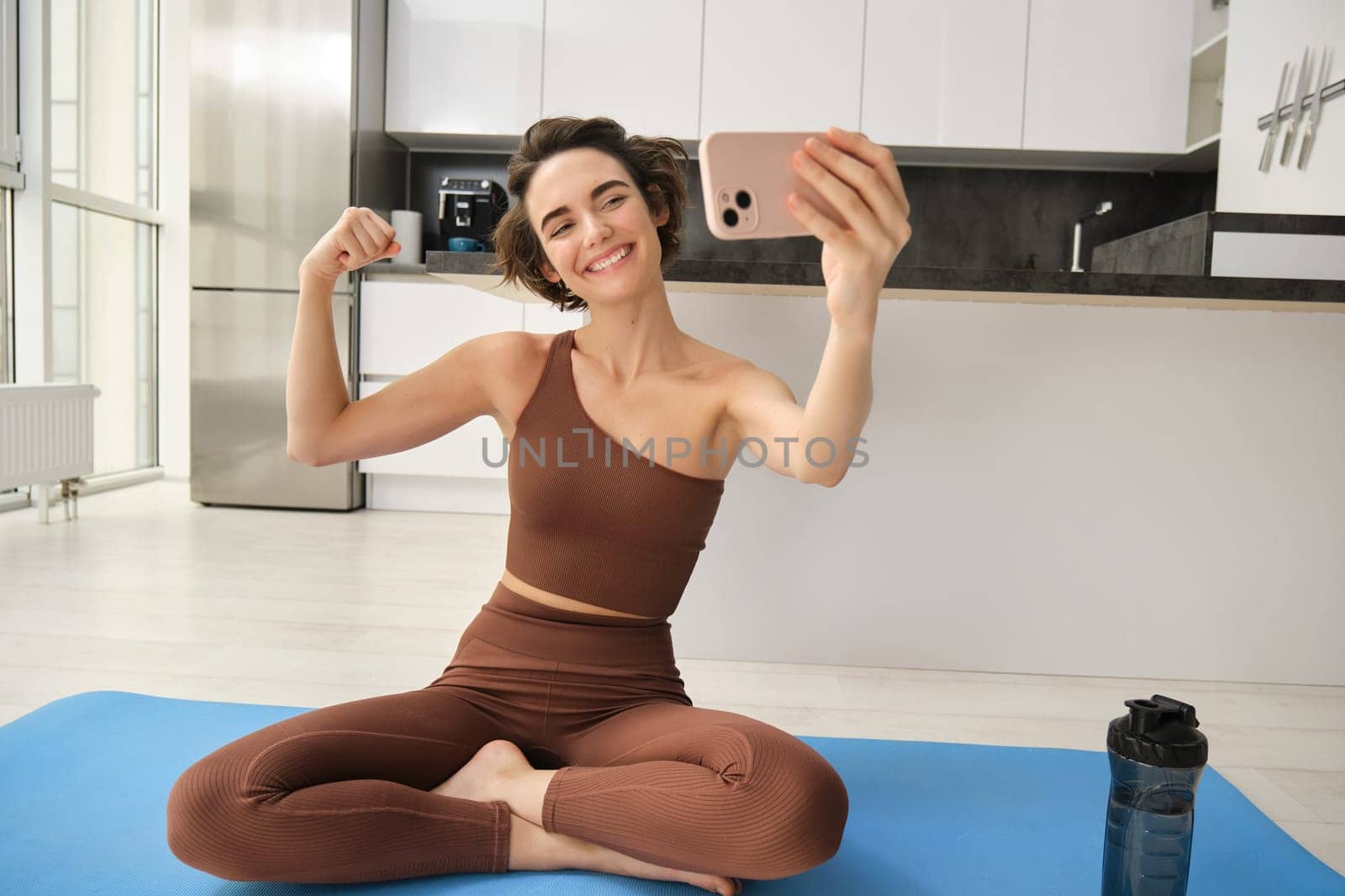 Fit and healthy woman fitness blogger, showing her muscles to smartphone camera, taking pictures in sportswear while doing yoga, workout at home and smiling.