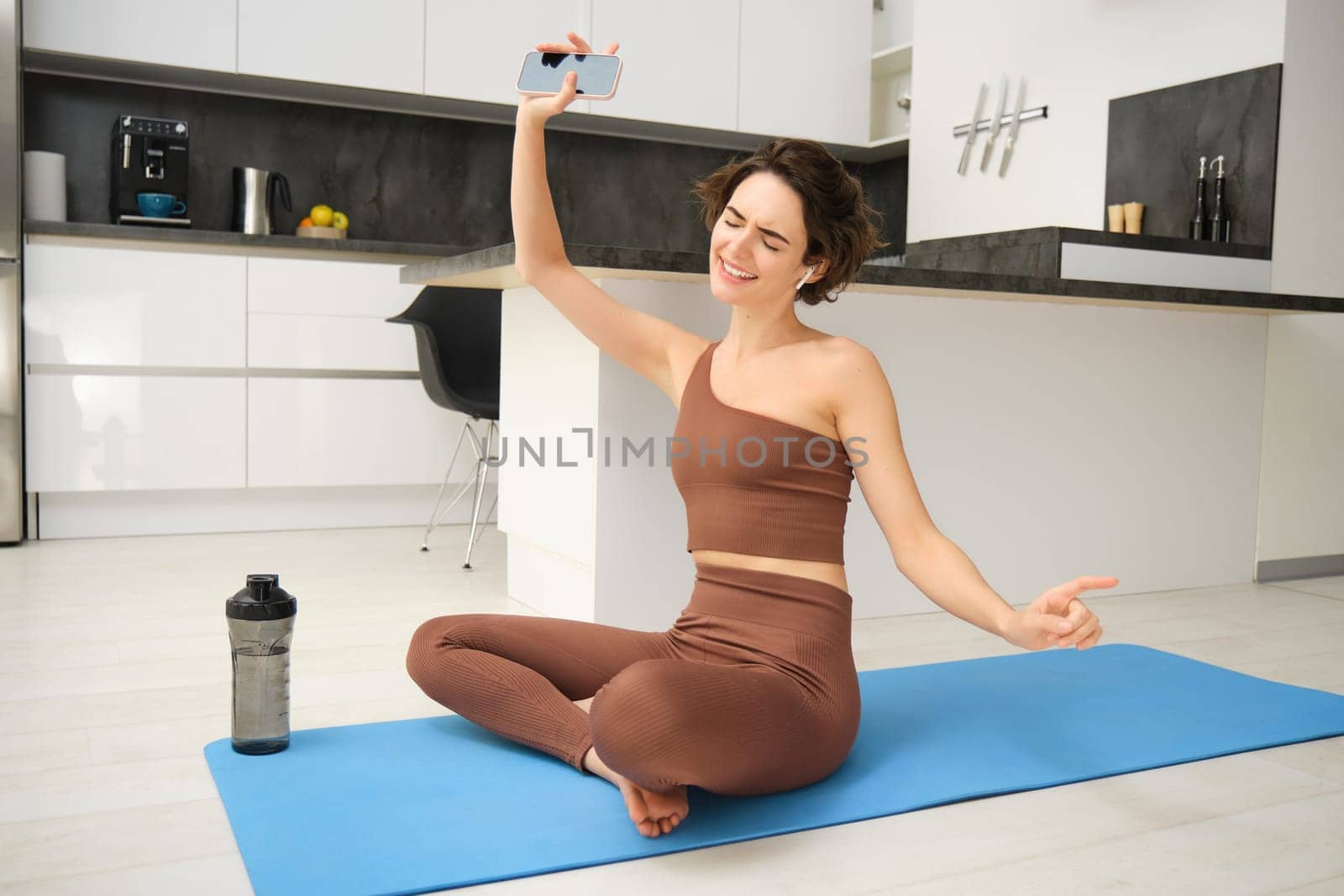 Happy brunette woman, sporty girl in activewear, dancing while sitting on yoga mat, listens music during sport workout at home.