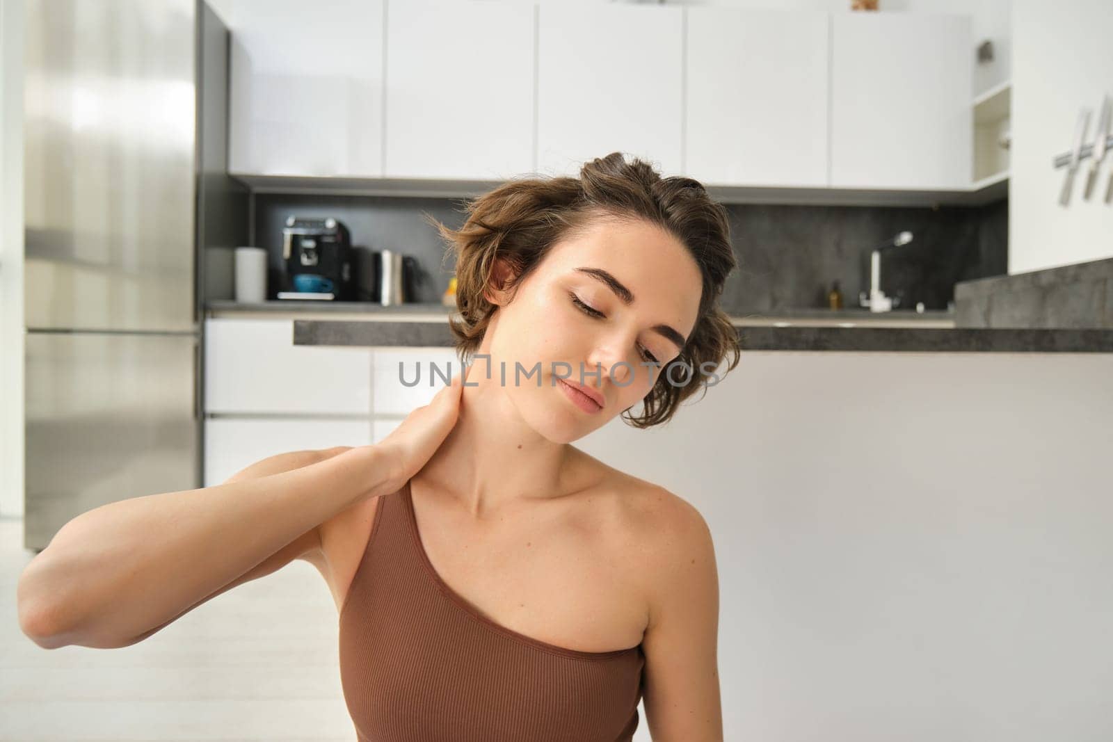 Fitness girl in activewear, doing yoga at home, stretching and working out, massaging her neck with pleased, relaxed face expression by Benzoix