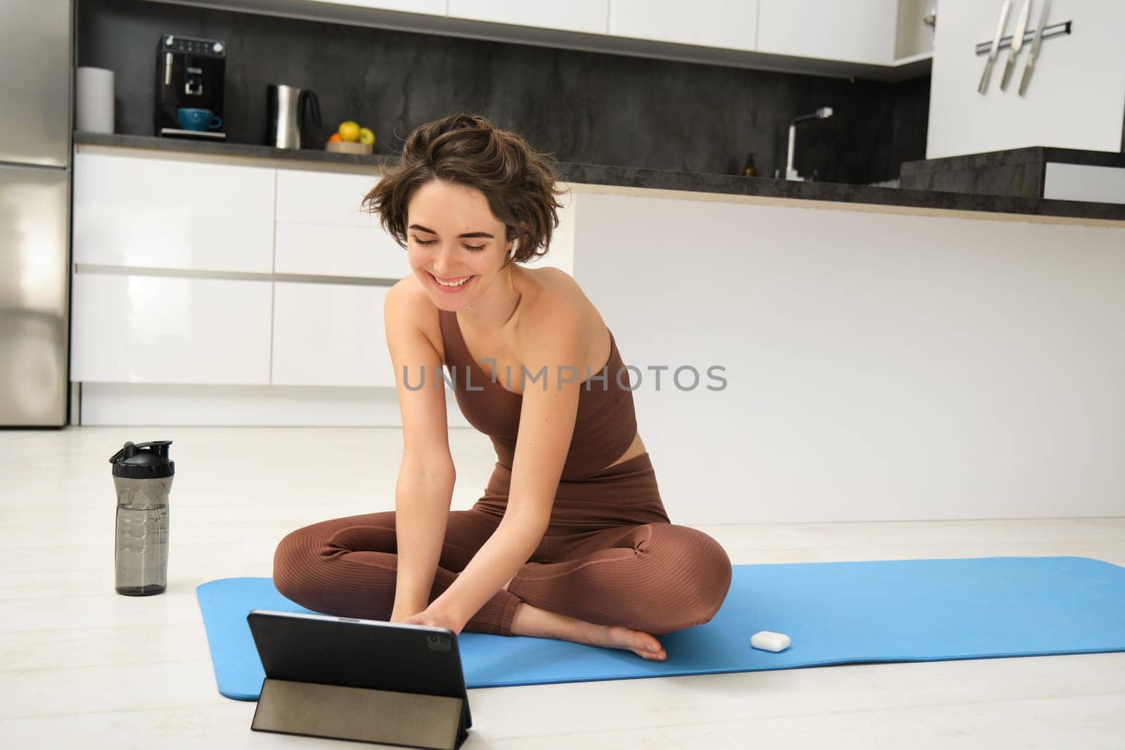 Sport and women health. Young brunette woman in fitness sportswear, sits on rubber mat, joins online workout class, remote yoga training on digital tablet, does stretching pilates exercises.