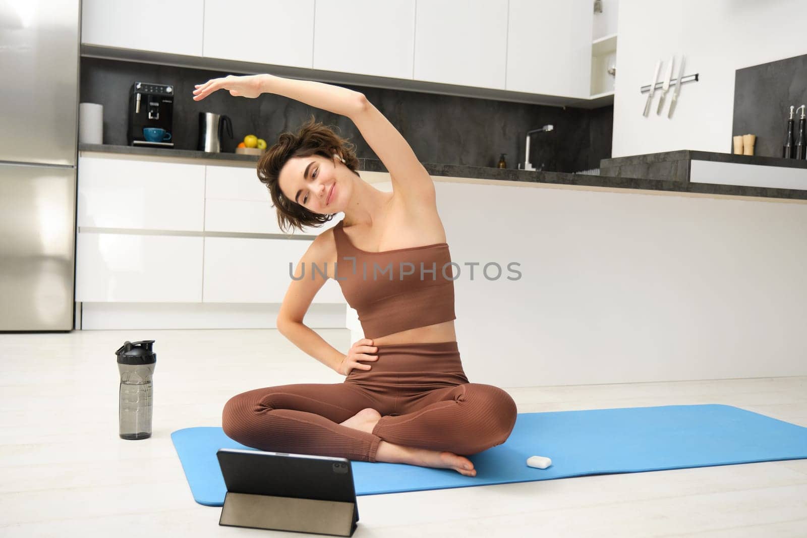 Young woman looking at digital tablet, connects to stretching fitness online class, workout, sits on yoga rubber mat at home, follows gym instructor exercises remotely by Benzoix