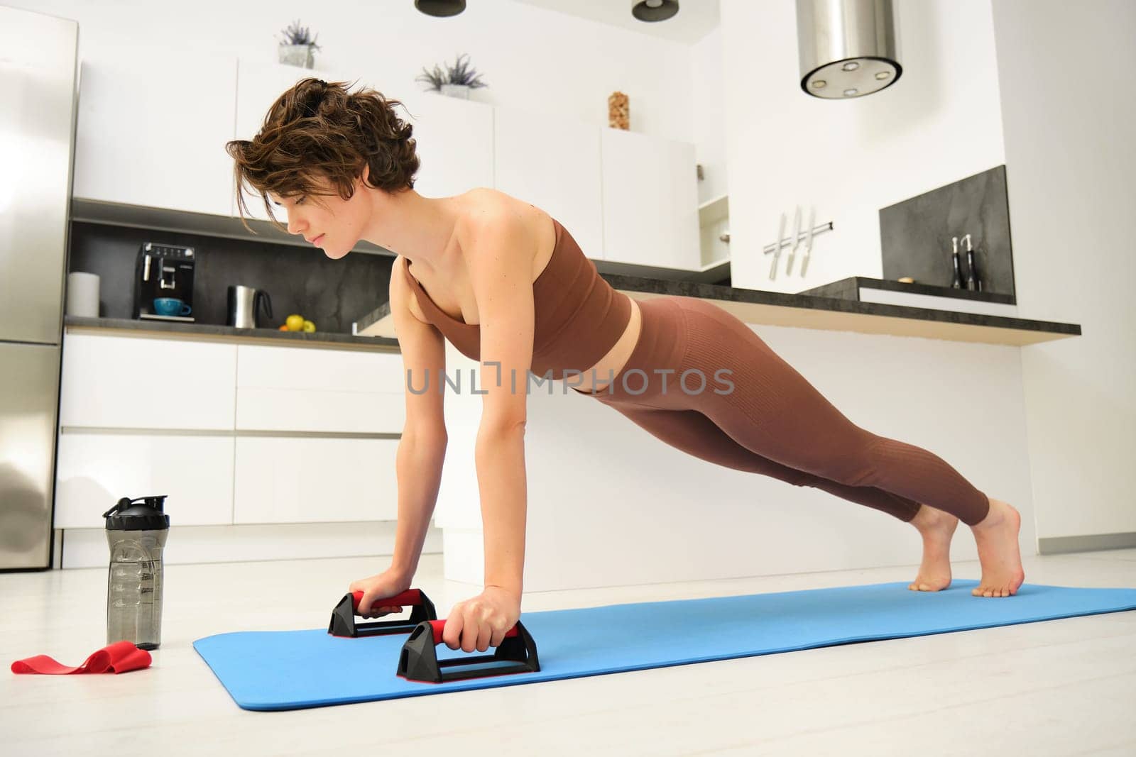 Portrait of young woman in sportswear, workout on yoga rubber mat, stands on push up bars stand for plunk exercises, fitness training session at home.