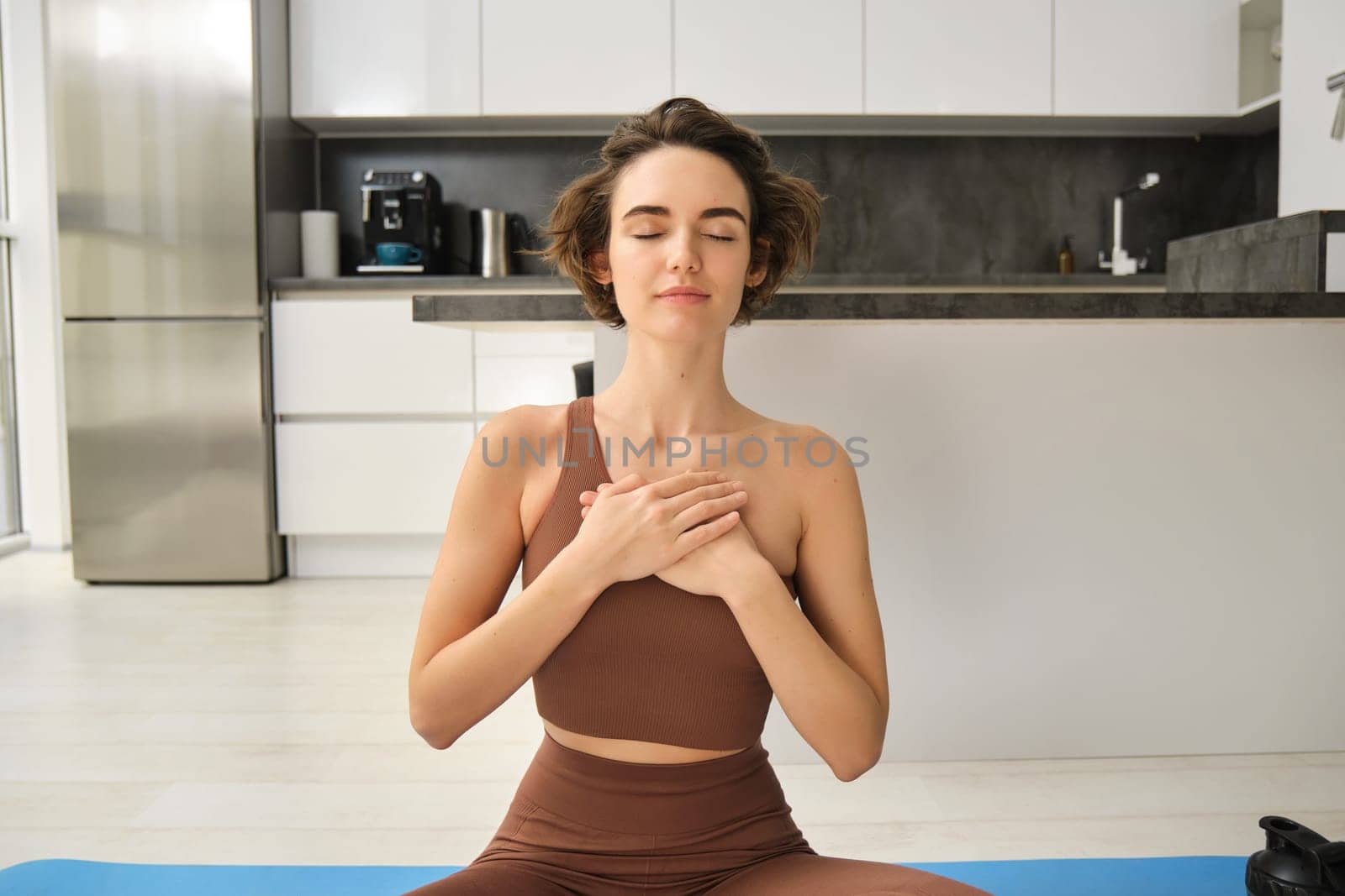 Heart chakra yoga. Portrait of woman practice mindfulness meditation at home, holding hands on chest with eyes closed and relaxed smile, sitting on rubber mat on kitchen floor by Benzoix