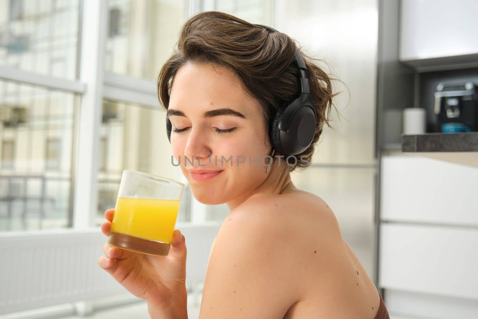 Sport and healthy lifestyle. Portrait of happy sporty girl, fitness woman in headphones, rewinds at home, drinks orange juice with happy smile after workout by Benzoix