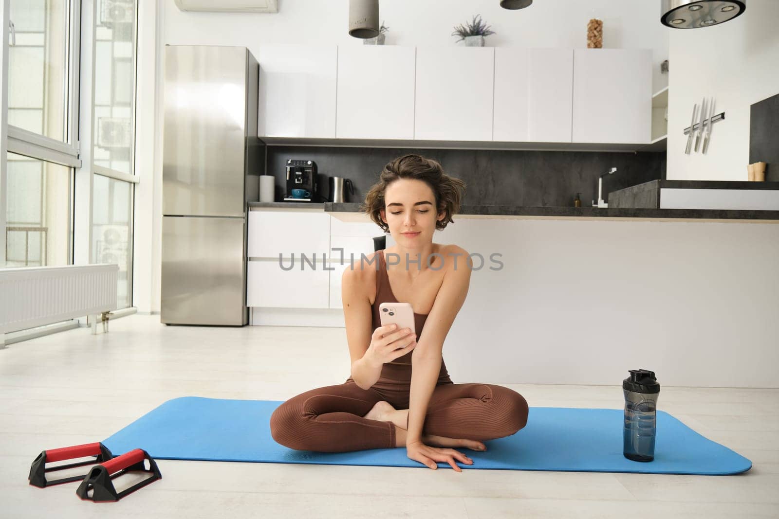 Yoga and sport at home concept. Young woman sitting in bright rome with mobile phone, doing workout on rubber mat, searching for exercises online on smartphone app, using sports equipment by Benzoix