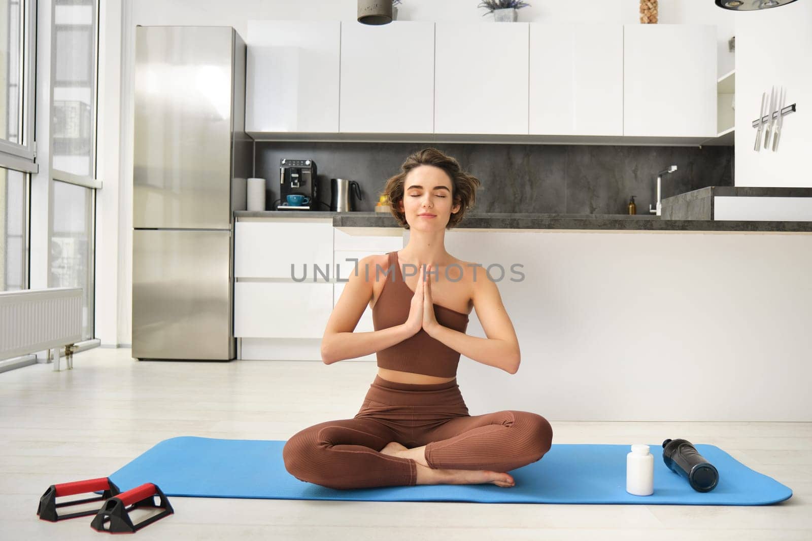 Smiling girl athlete, sportswoman doing yoga at home in activewear, sitting on yoga rubber mat in lotus pose, meditating, practice mindfulness exercises.