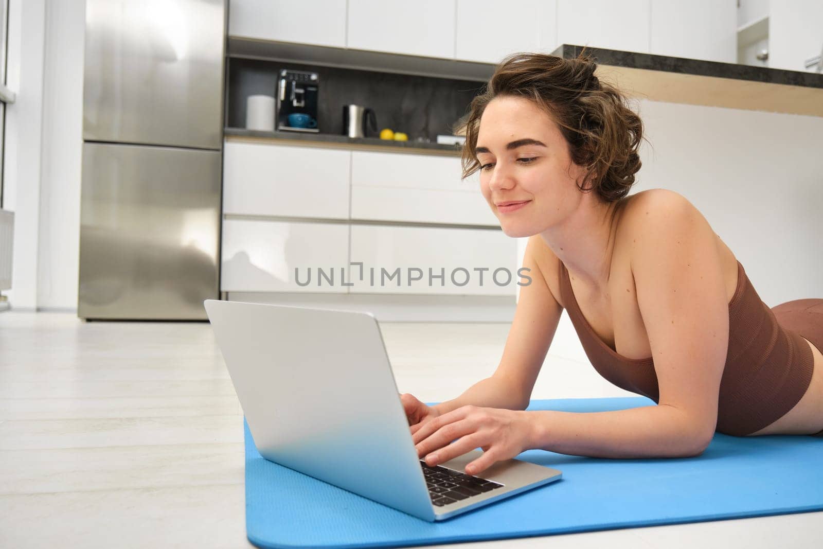 Portrait of young healthy woman in sportswear, lying on yoga mat, looking for fitness videos on laptop, workout from home. Sport and wellbeing concept