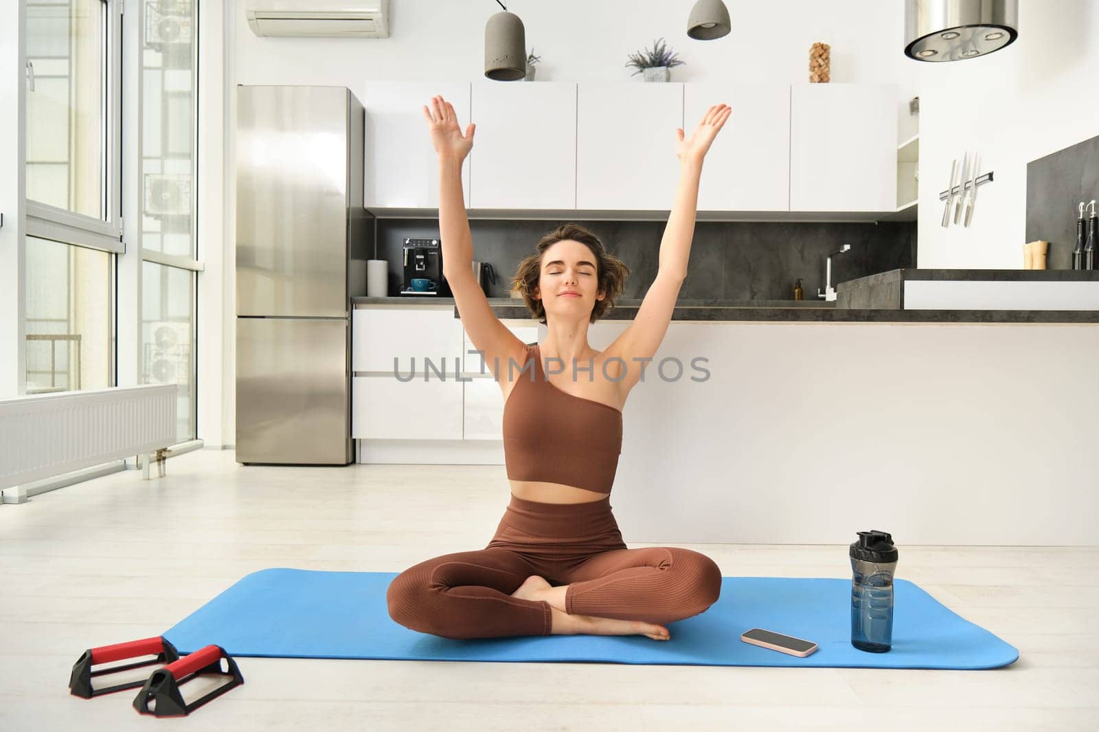 Smiling young woman in activewear, sitting on mat at home, raising arms up while meditating, practice yoga, doing mindful workout by Benzoix