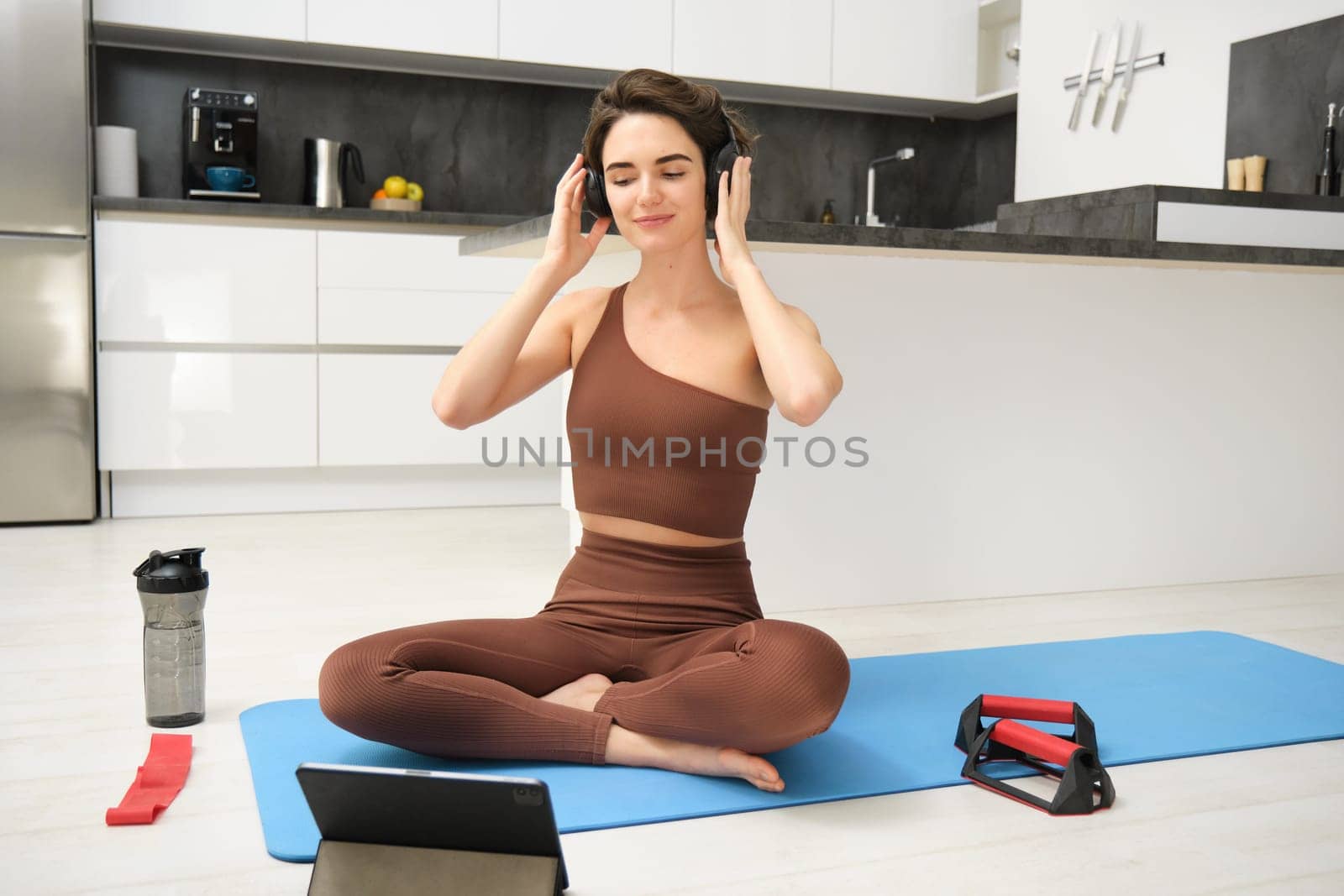 Smiling fitness girl in headphones, watches pilates, gym instructor online video, workout at home on yoga rubber mat, uses plank equipment and resistance band.