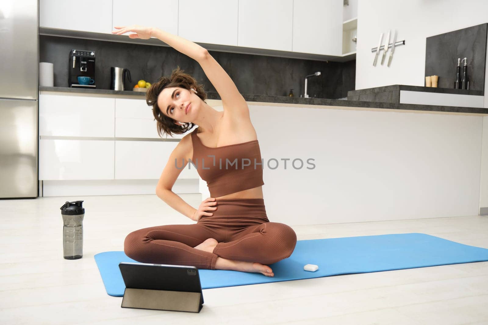 Young woman looking at digital tablet, connects to stretching fitness online class, workout, sits on yoga rubber mat at home, follows gym instructor exercises remotely.