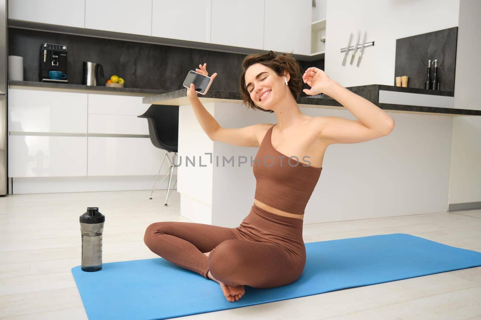 Active lifestyle and health. Happy young woman does workout, sits and dances with smartphone in hands, listens music in wireless earphones during fitness training session, yoga on floor mat.