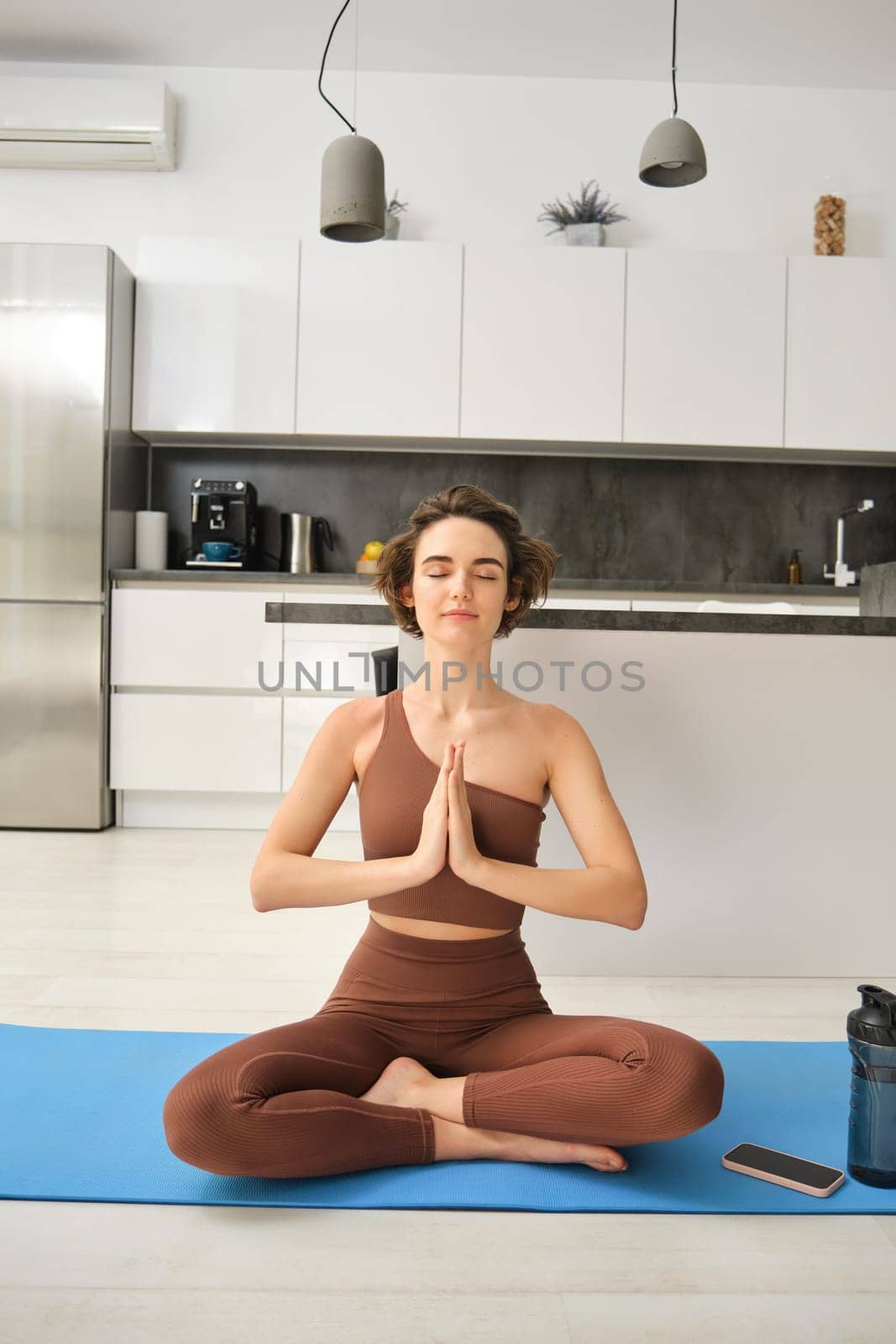 Vertical shot of young woman meditating, doing yoga at home on rubber mat at home.