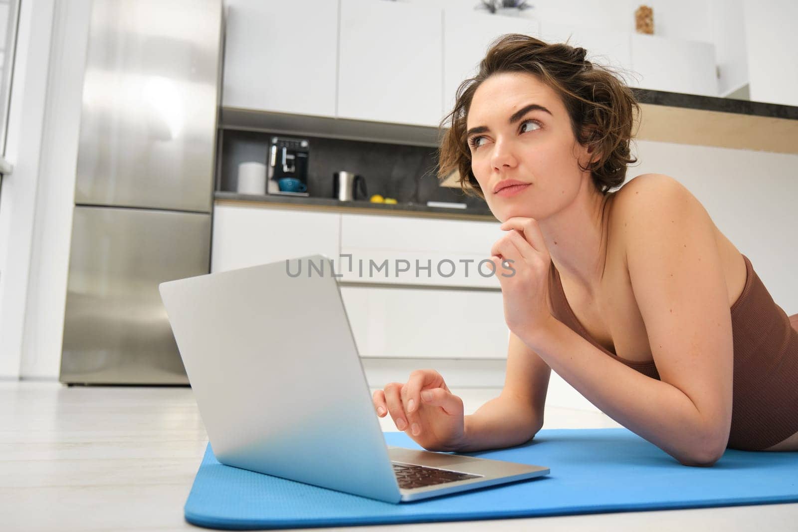 Fitness girl lying on rubber mat with thoughtful face, using laptop, fitness instructor thinking, planning online class, workout at home in kitchen by Benzoix