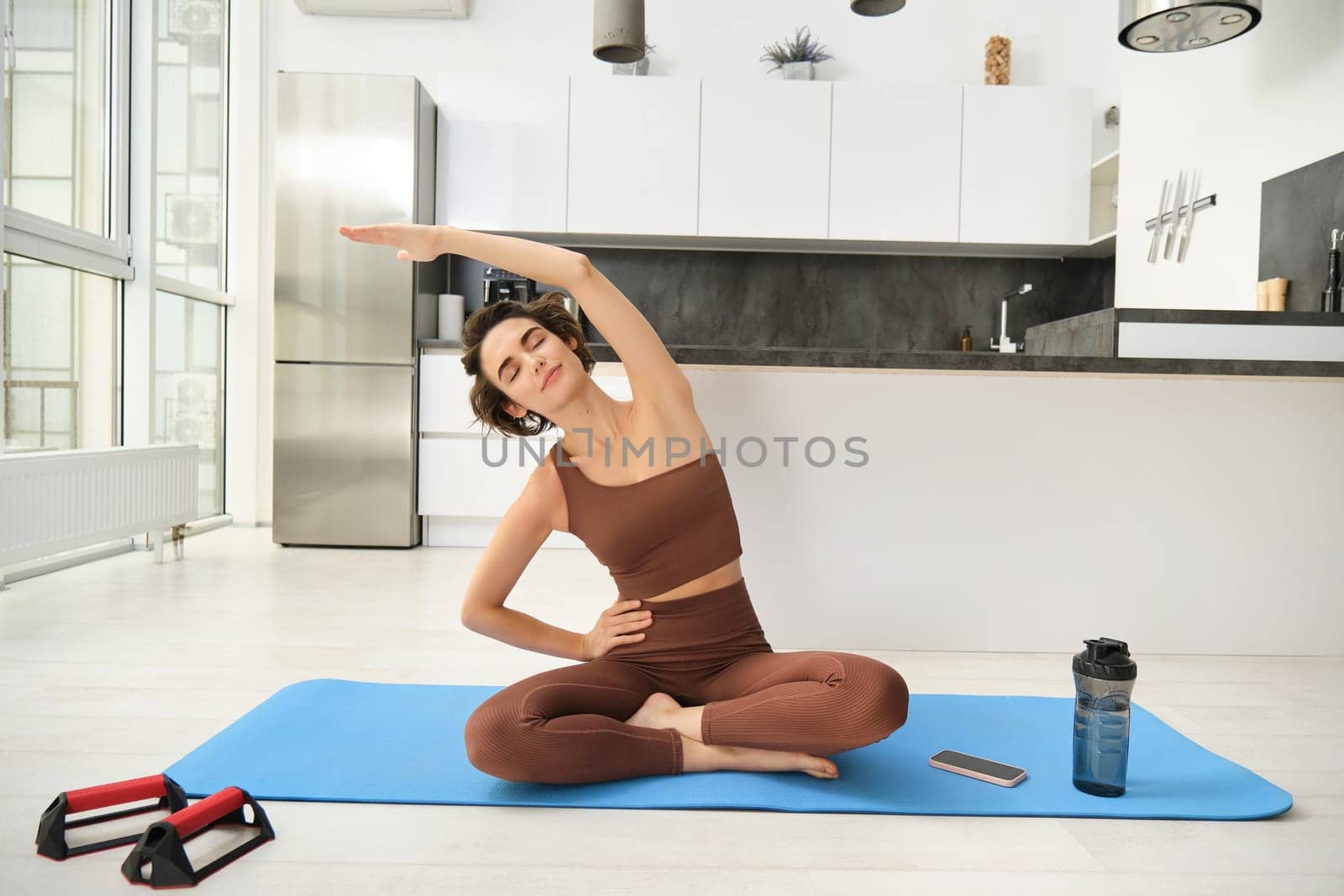 Fitness girl doing exercises at home. Young woman athlete sitting on yoga mat in bright room, stretching her arms, doing pilates exercises by Benzoix
